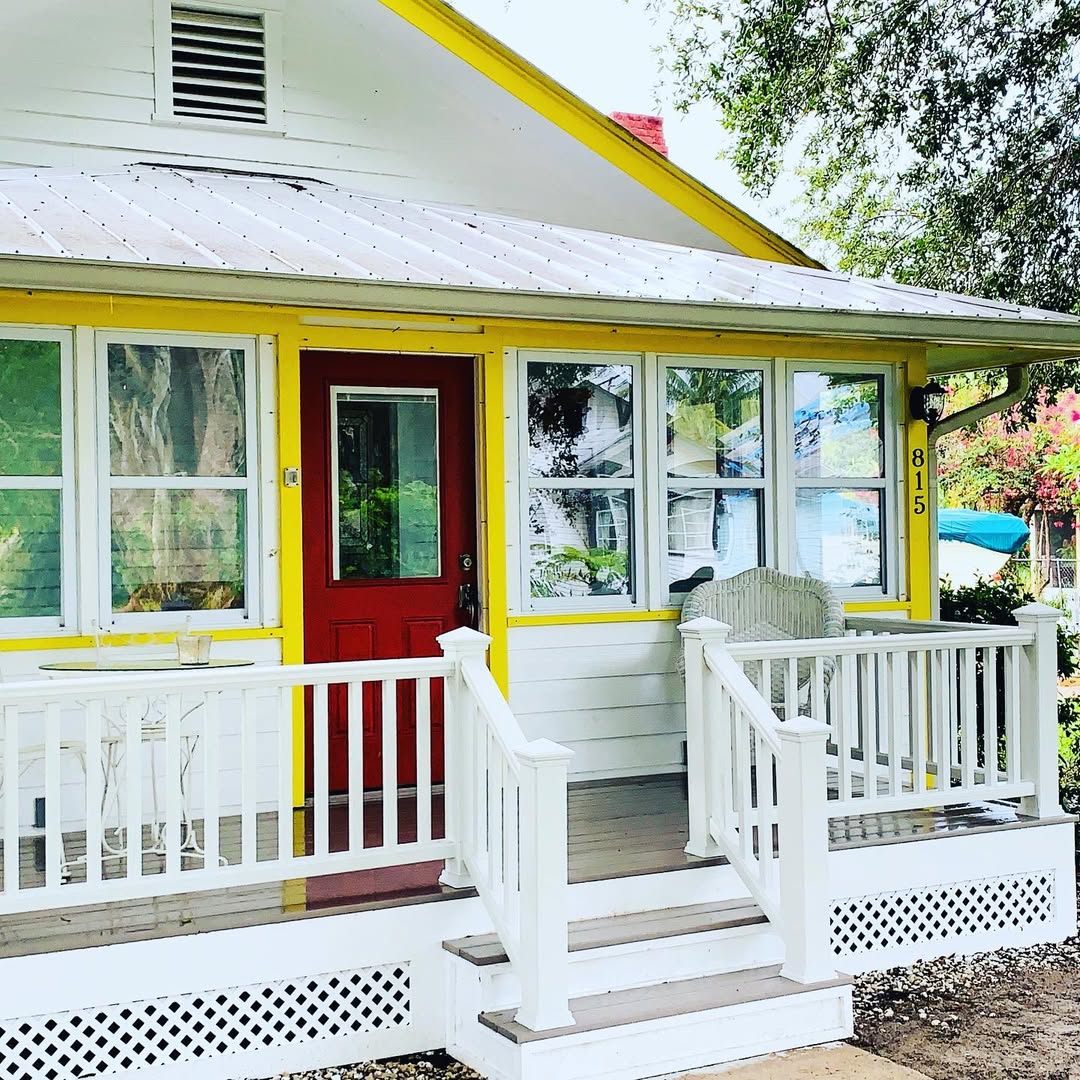 A small white house with yellow trim, a red front door, covered porch, and white railings showcases Coastal Cottage Exterior Design—an inviting space that reflects 2025 Exterior Trends. A chair by the entrance welcomes you to this home, numbered 815.