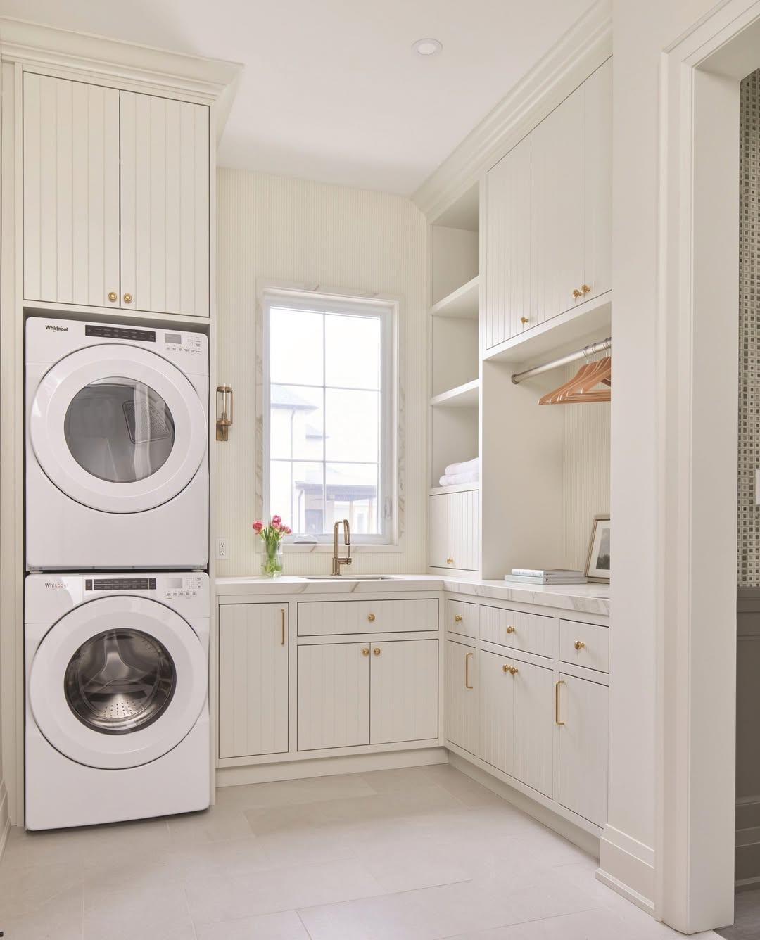 A bright stacked laundry room features a washer and dryer, built-in cabinets, countertop, sink, window, and a hanging rod with wooden hangers for practical organization.