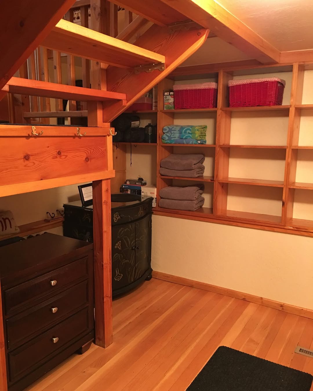 A wooden staircase with open shelves holding towels, baskets, and various items showcases clever under stairs closet ideas; a dark dresser and cabinet are placed beneath the stairs on a hardwood floor.