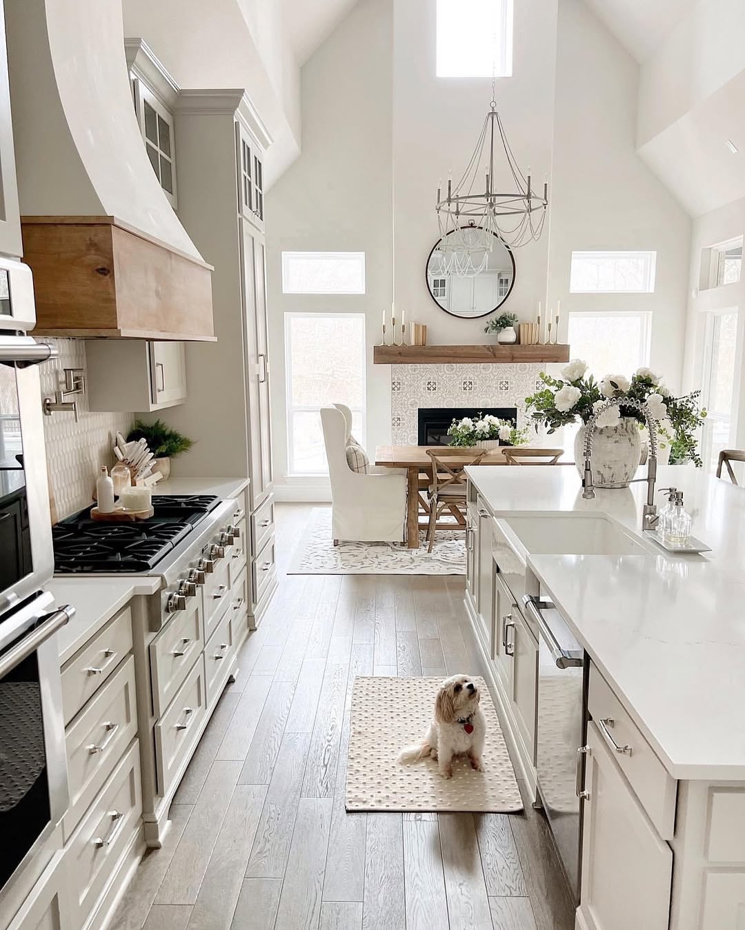 A modern white farmhouse kitchen with a center island, stainless steel appliances, large windows, and a dining area with a round table—perfect for those seeking fresh farmhouse kitchen ideas. A small dog relaxes on a rug atop the wooden floor.