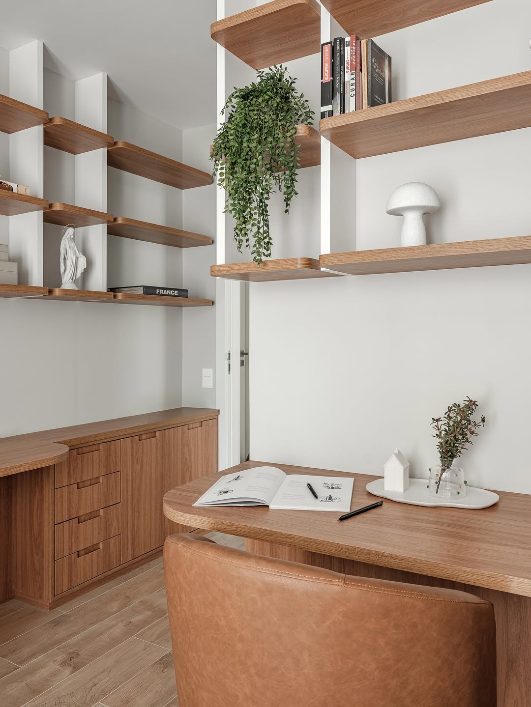 Modern home office with wooden shelves, a leather chair, a desk holding an open book, a pen, a small vase with greenery, and stylish decor—perfect inspiration for your next home office ideas.