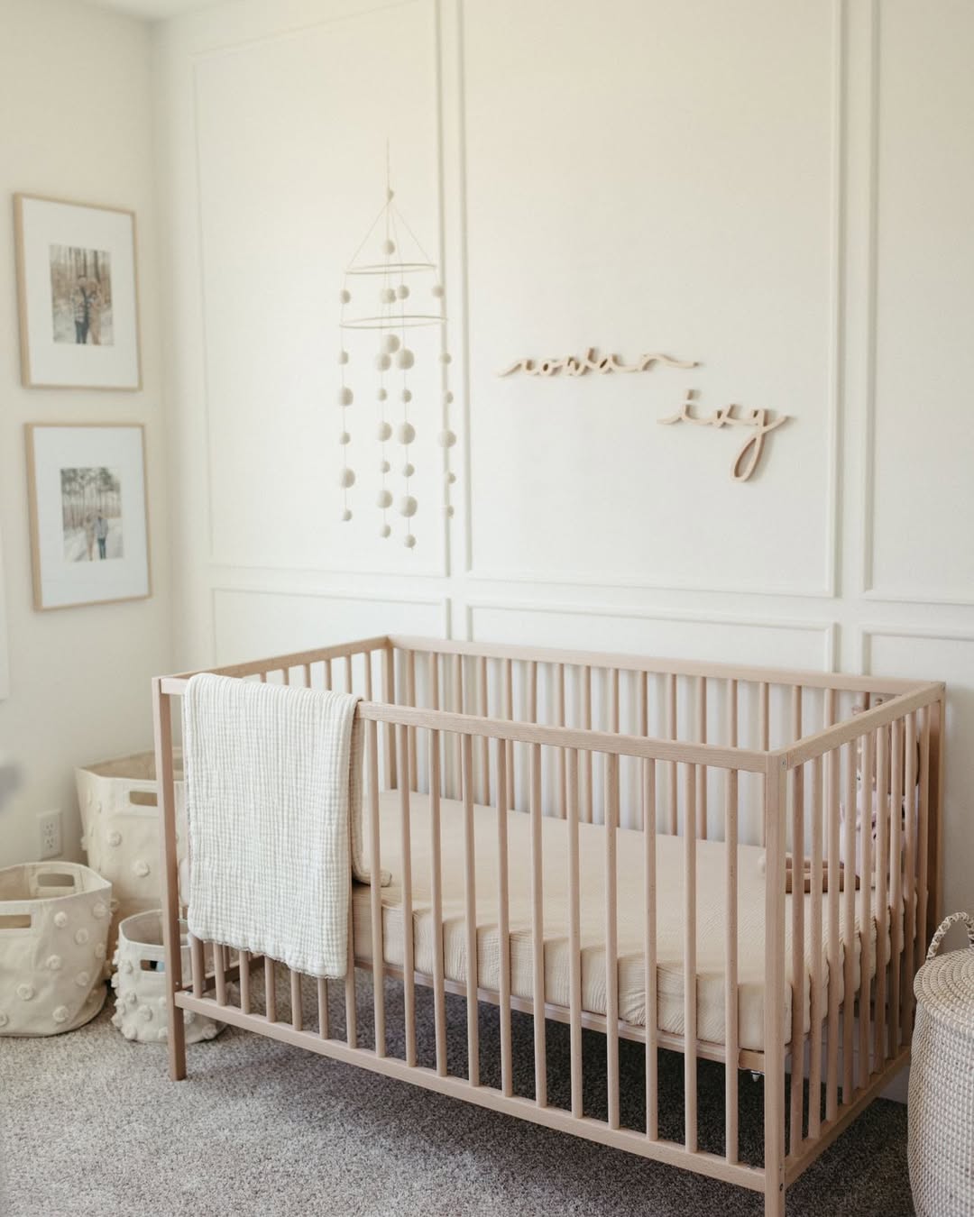 A minimal nursery with a wooden crib, cream bedding, textured blanket, wall art, a hanging mobile, framed photos, and cream storage baskets—perfect baby boy nursery ideas for the first-time mom in 2025.