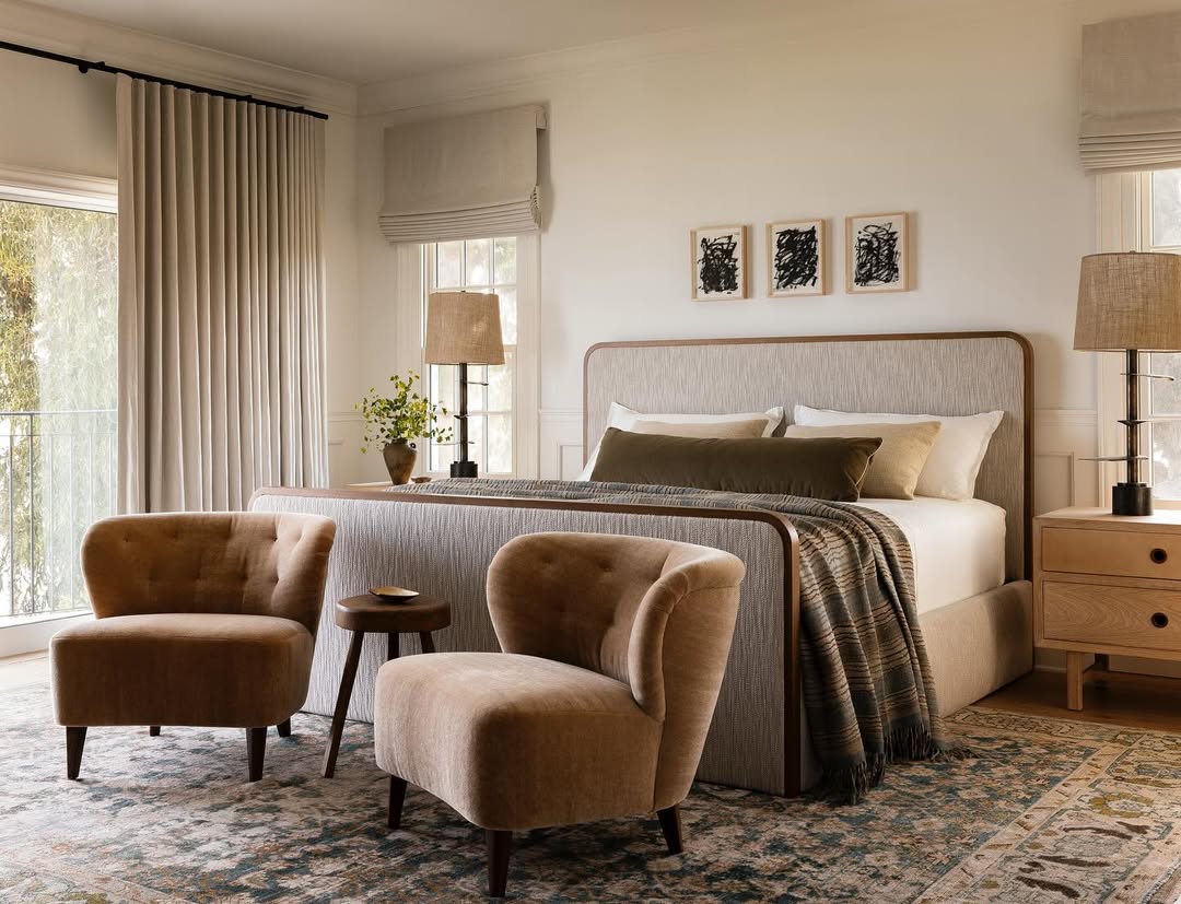 A modern bedroom with a neutral color palette, perfect for interior lovers seeking 2025-inspired bedroom color schemes, featuring a bed, two upholstered chairs, a small round table, lamps, nightstands, and framed art above the headboard.