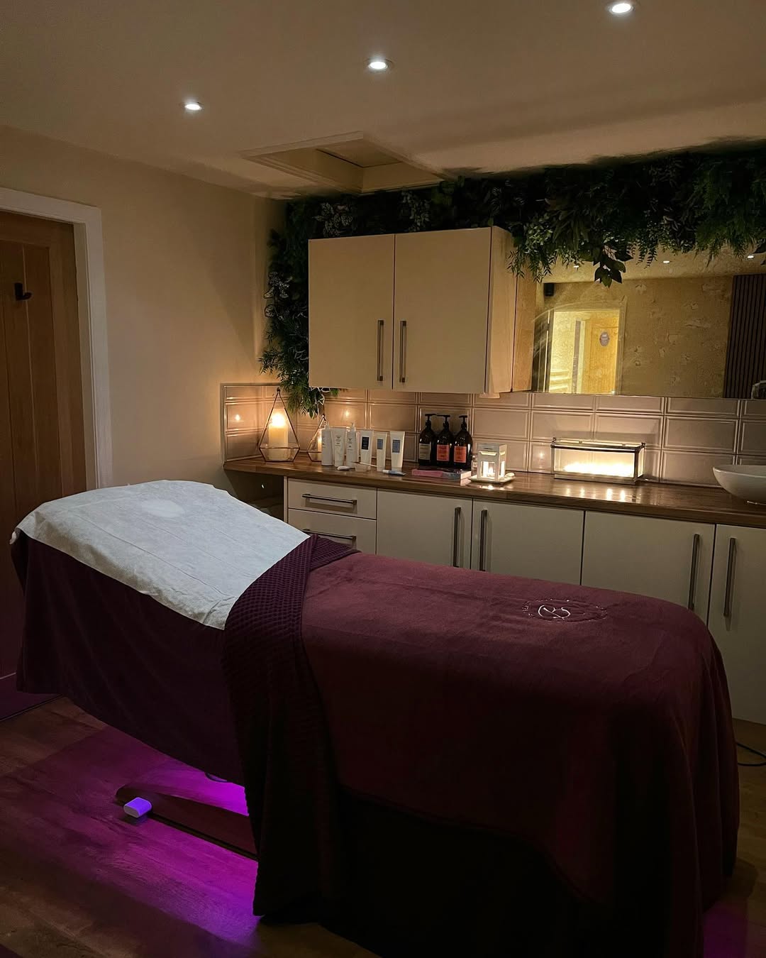 A massage room with a burgundy blanket on the table; cabinets, candles, bottles, and soft lighting offer inspiring massage room ideas for a truly relaxing atmosphere.