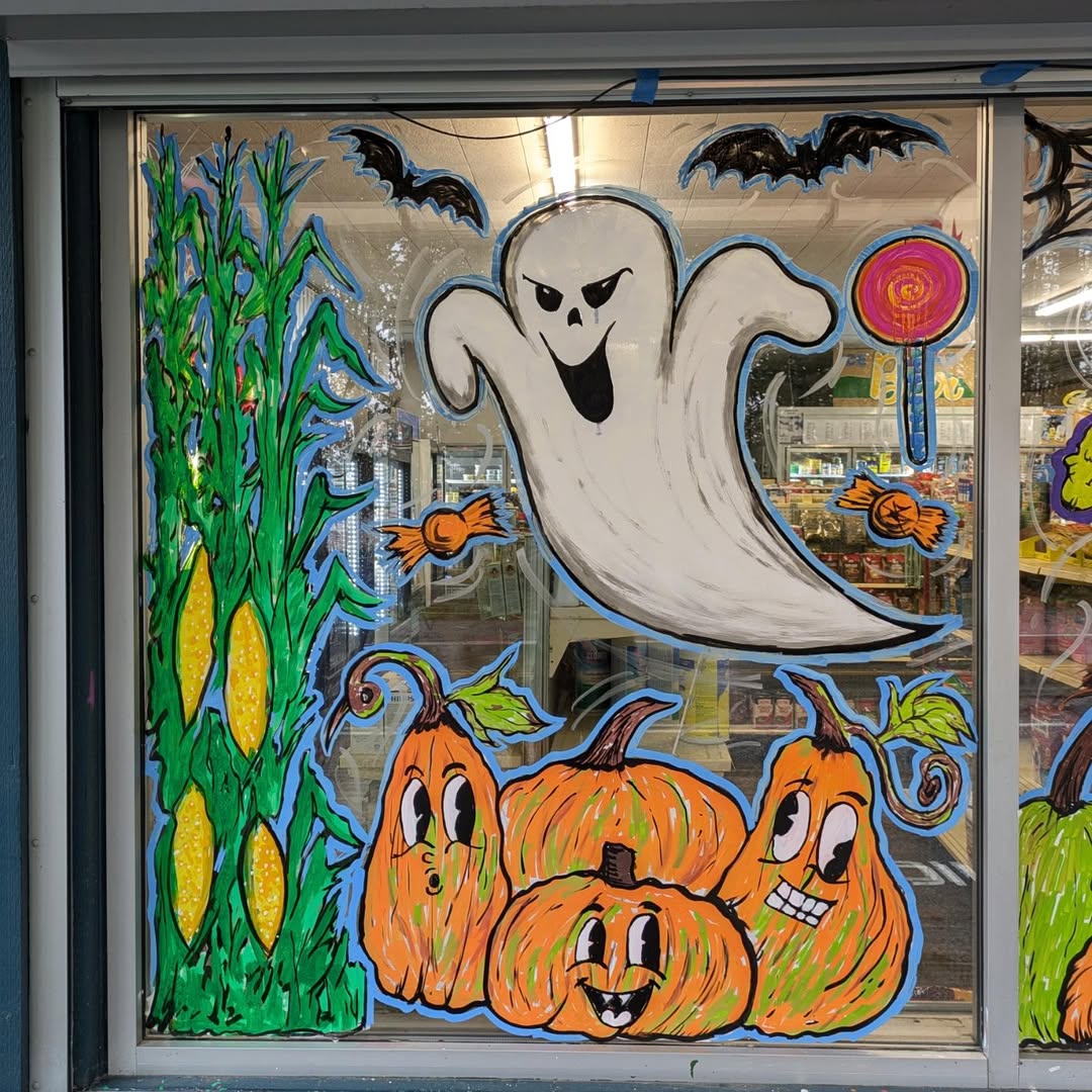A store window painted with a ghost, three pumpkins with faces, corn stalks, bats, candy, and a lollipop offers festive Halloween decorations and great Halloween window decoration ideas.