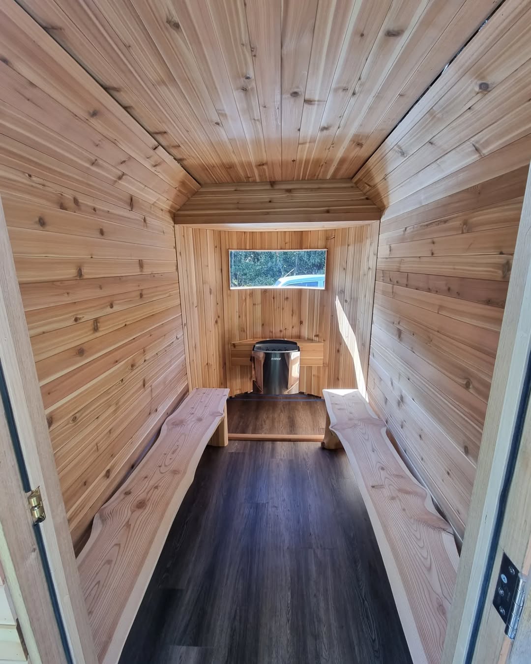 A small wooden sauna interior with two benches along the walls, a heater at the far end, and natural light streaming through a rectangular window—perfect inspiration for cozy outdoor sauna ideas.