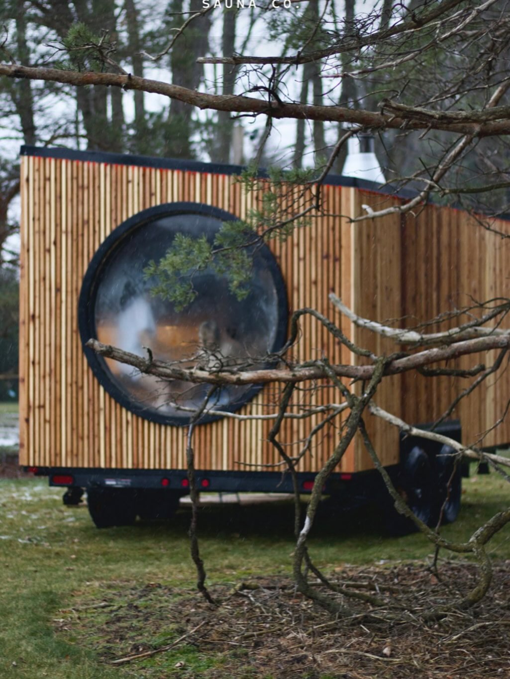 A wooden mobile sauna with a large circular window sits on a grassy area, partially obscured by tree branches in the foreground—an inspiring example of gorgeous outdoor saunas and innovative sauna design.