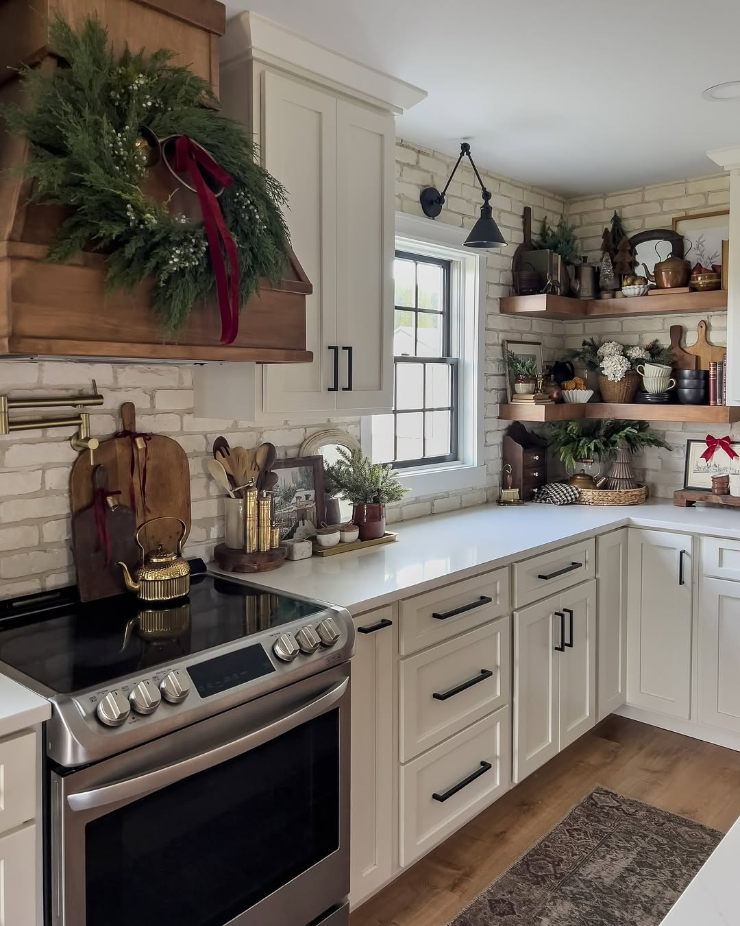 A charming cottagecore kitchen with white cabinets, wooden shelves holding plants and decor, a stove with a kettle, and a vent hood adorned with greenery and a red ribbon—perfect inspiration for cottagecore decor kitchen ideas.