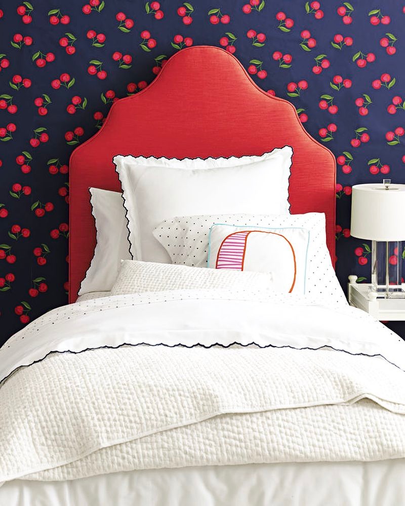 A bed with a red headboard, white bedding, and decorative pillows sits against a navy blue wall with a red cherry pattern—perfect for those seeking unique Cherry Bedroom Ideas. A white lamp completes the stylish bedside table setup.