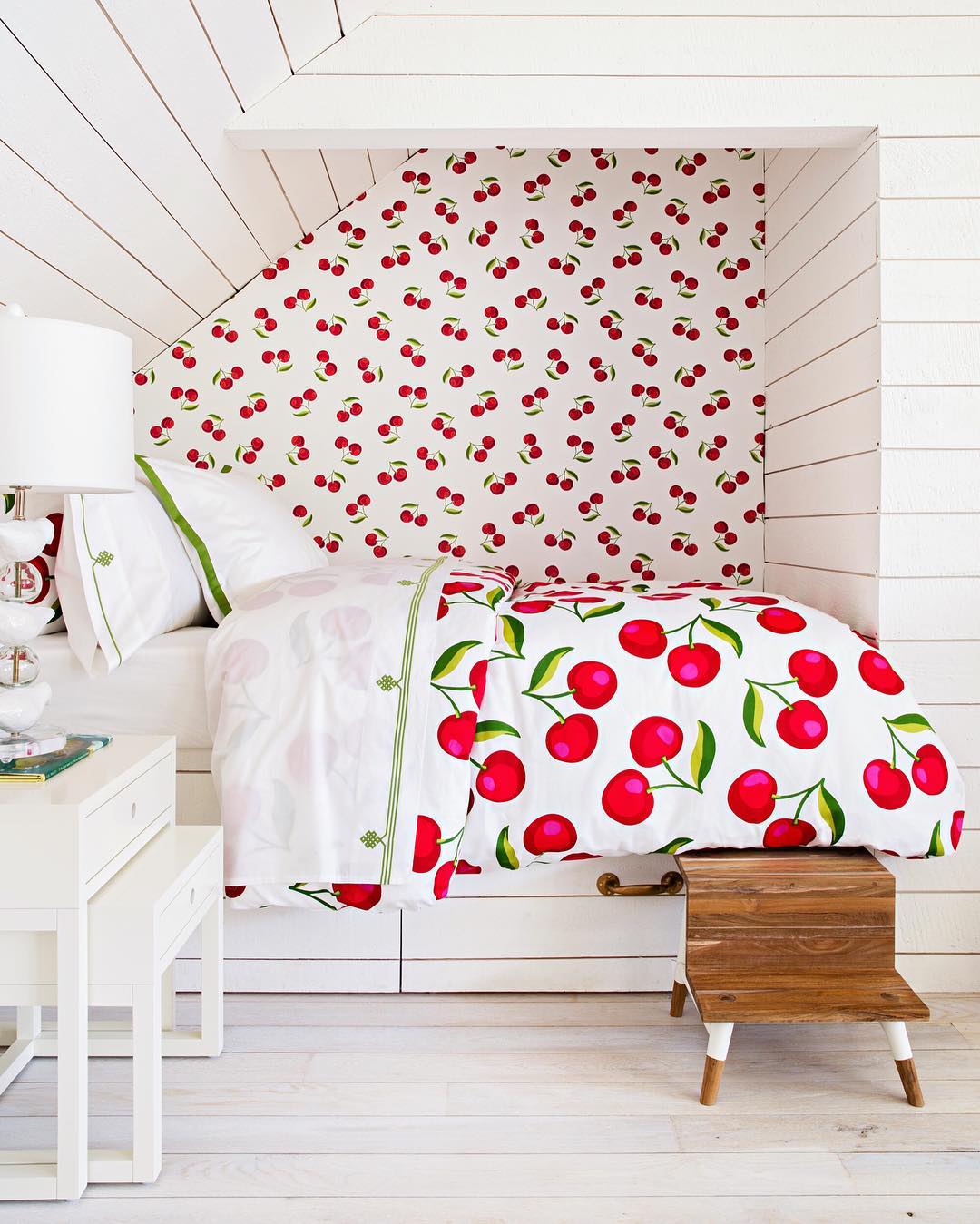 A bed with cherry-patterned bedding and matching wallpaper is set in a white-paneled nook, offering fresh Cherry Bedroom Ideas. A nightstand with a lamp and a small wooden step stool are beside the bed.