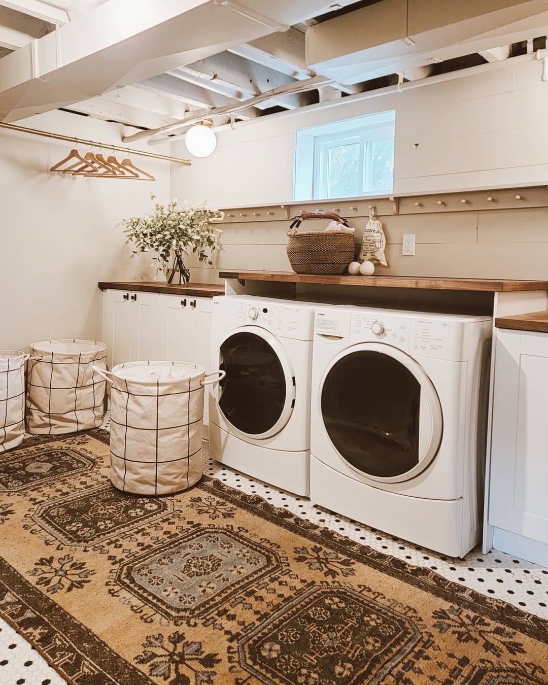 A boho laundry room featuring a front-loading washer and dryer, white cabinets, wire baskets, hanging rods with hangers, and a patterned rug on a tiled floor—perfect inspiration for unique laundry room ideas.