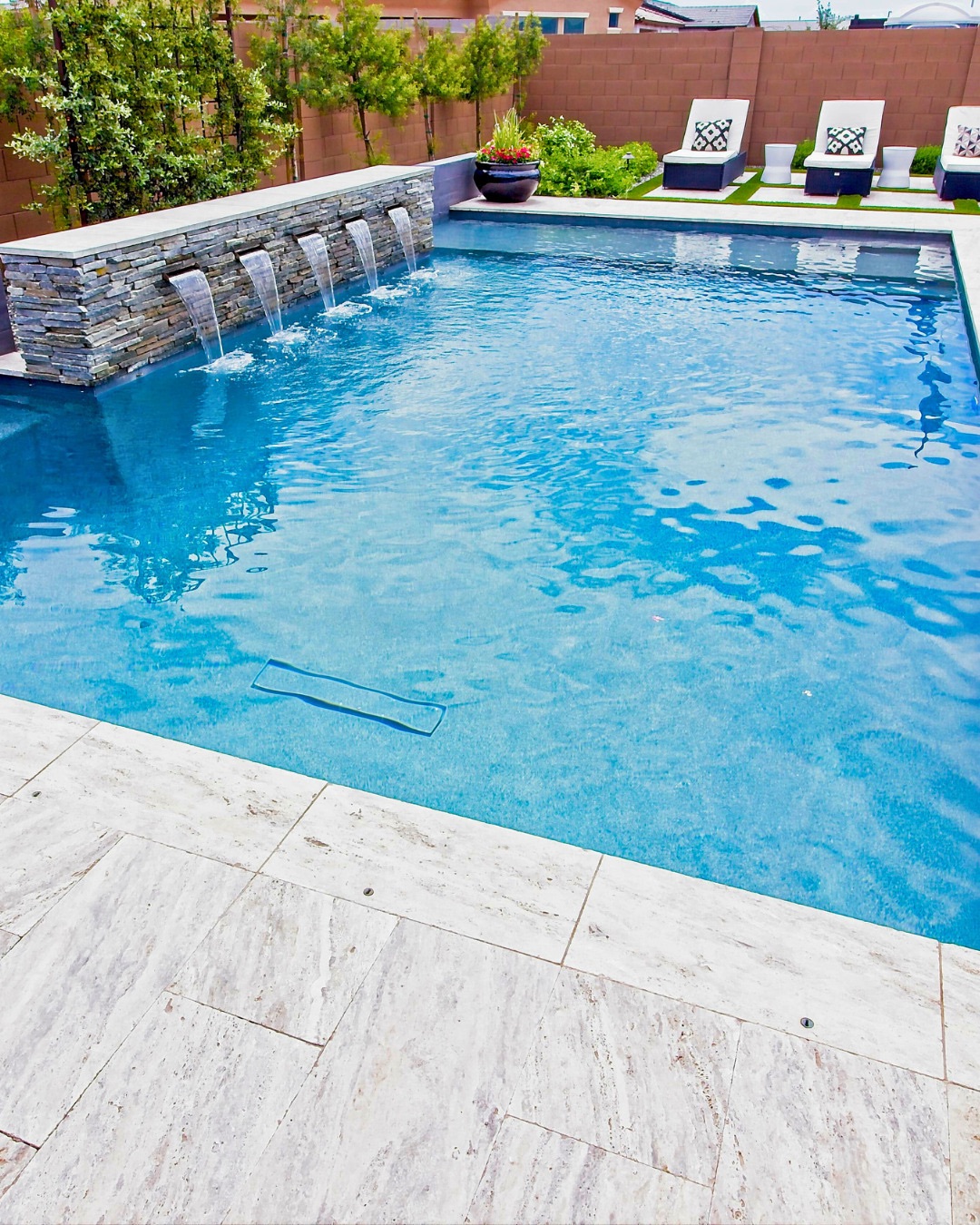 Rectangular swimming pool with three water features on a stone wall, surrounded by paved tiles and poolside lounge chairs with pillows—a stunning example of cool pool ideas for stylish backyard pools.