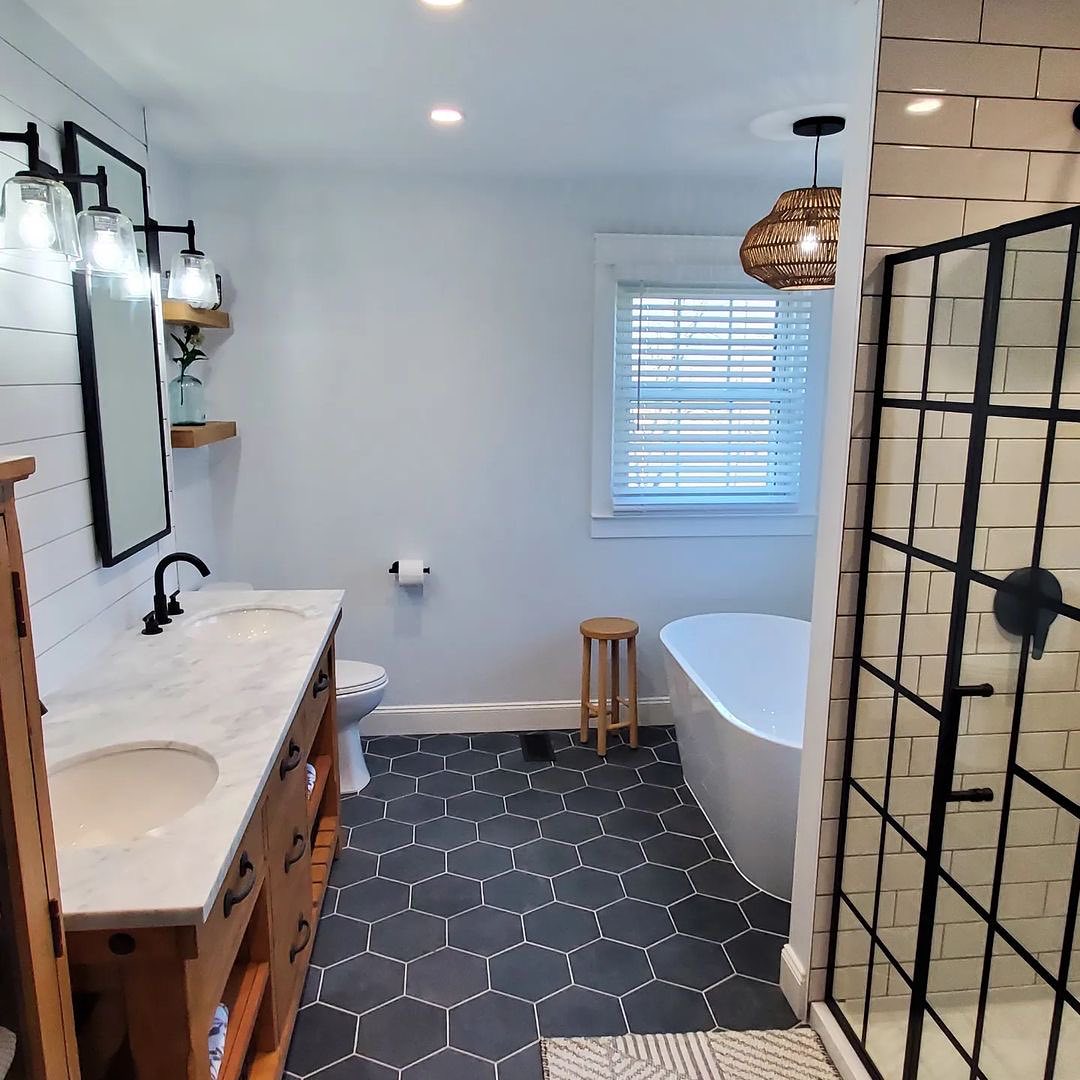 Modern bathroom featuring black hexagonal floor tiles—perfect for inspired bathroom tile ideas—a double sink vanity, freestanding bathtub, glass shower enclosure, and a window with white blinds for a spa-worthy bathroom design.