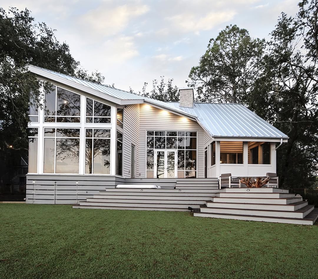 Ultra-stylish modern house with large floor-to-ceiling windows, a slanted roof, spacious wooden deck, and green lawn surrounded by trees—perfect for those seeking design ideas 2025 with a hint of coastal cottage exterior.