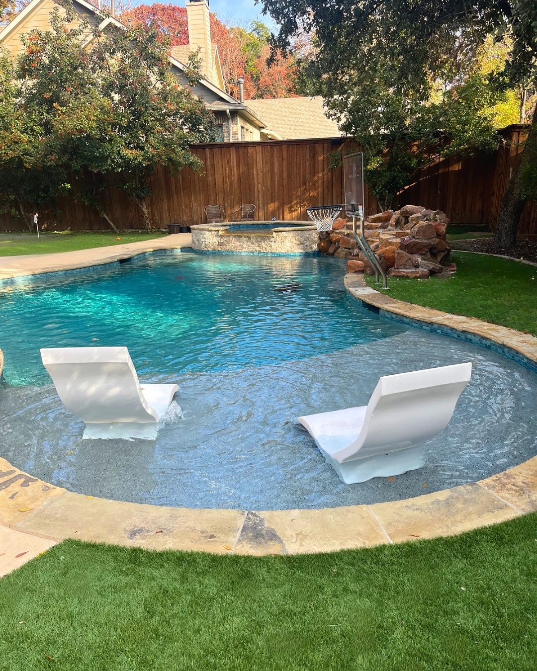 A backyard swimming pool with two white lounge chairs in the shallow area, a basketball hoop, and a stone waterfall feature—perfect inspiration for cool pool ideas—surrounded by lush grass and trees.