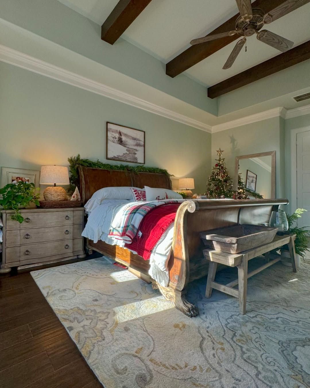 A sunlit sage green farmhouse bedroom with a wooden sleigh bed, Christmas decorations, a bench at the foot of the bed, a nightstand, and a large area rug.
