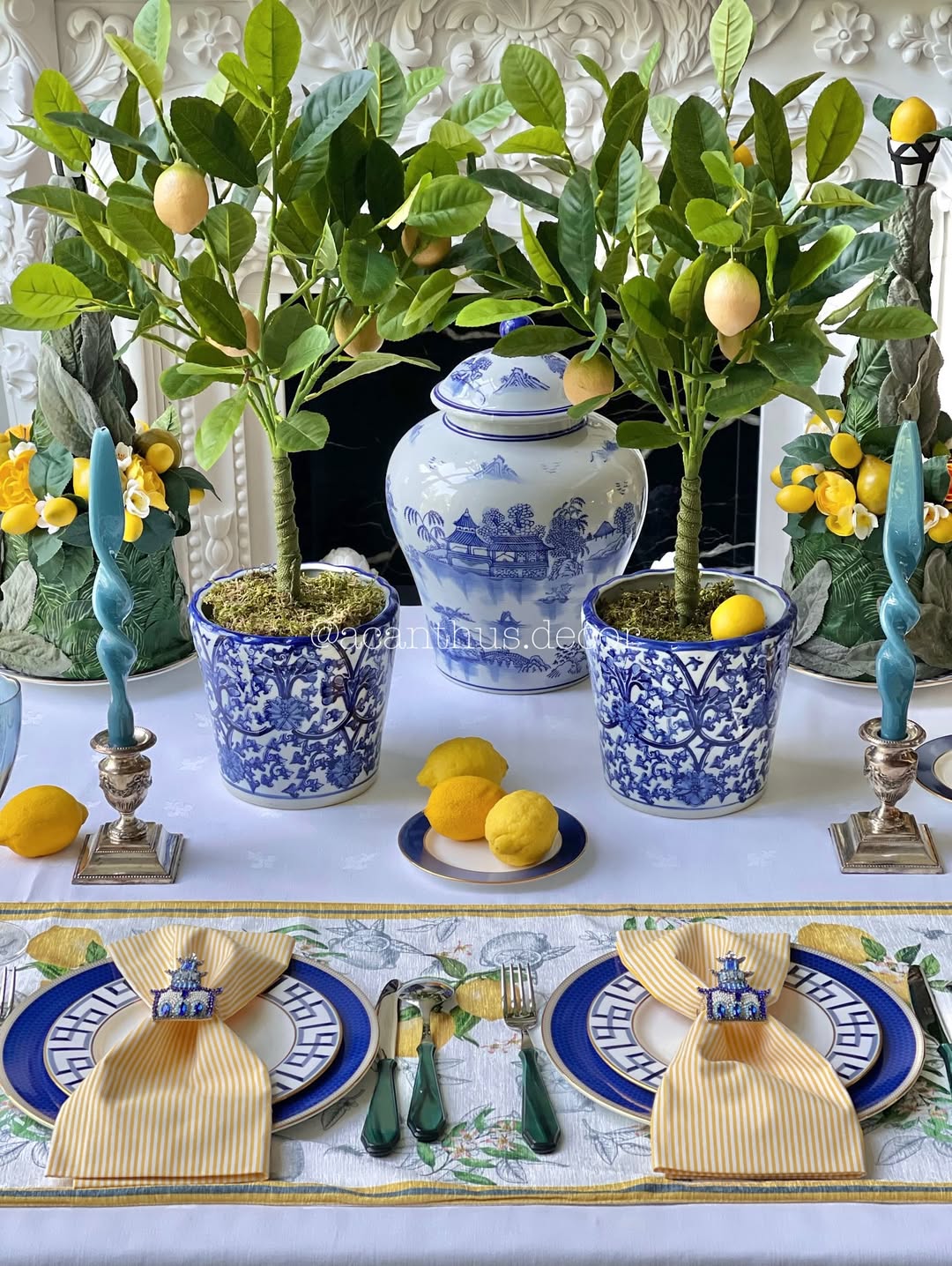 A dining table is set with blue and white plates, yellow napkins, green cutlery, and decorated with potted lemon trees, whole lemons, and a blue and white porcelain jar as the centerpiece—perfect for fresh table setting ideas.