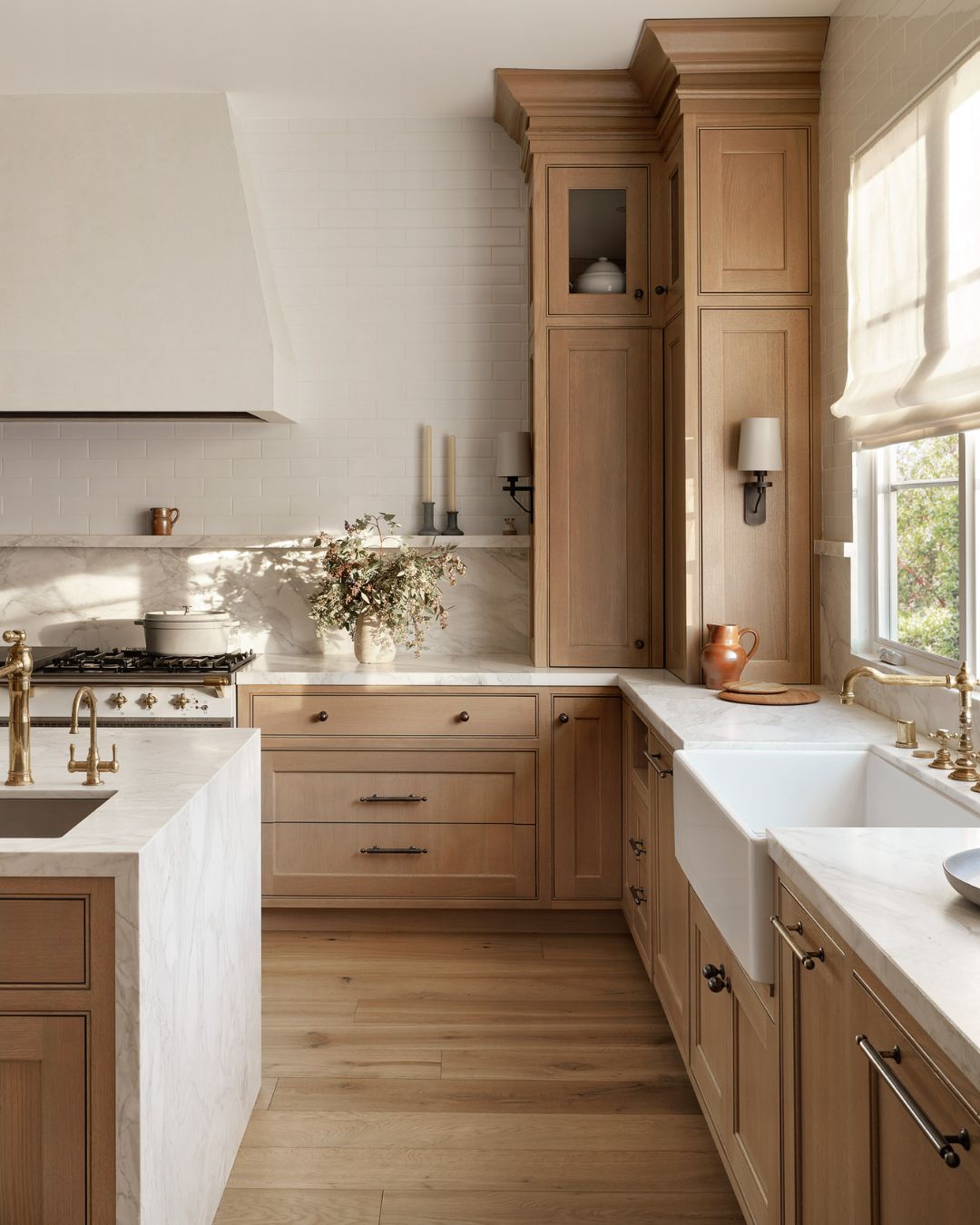 A modern kitchen design featuring light oak cabinets, white marble countertops, a farmhouse sink, brass fixtures, and natural light streaming through a window with a sheer curtain.