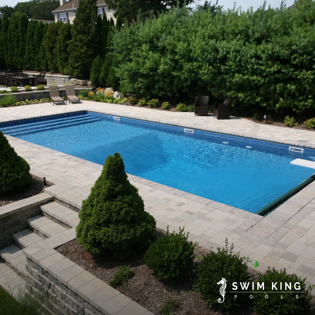 Rectangular inground swimming pool surrounded by stone pavers, landscaping, and lounge chairs—perfect inspiration for cool pool ideas and backyard pools; Swim King Pools logo in the corner.