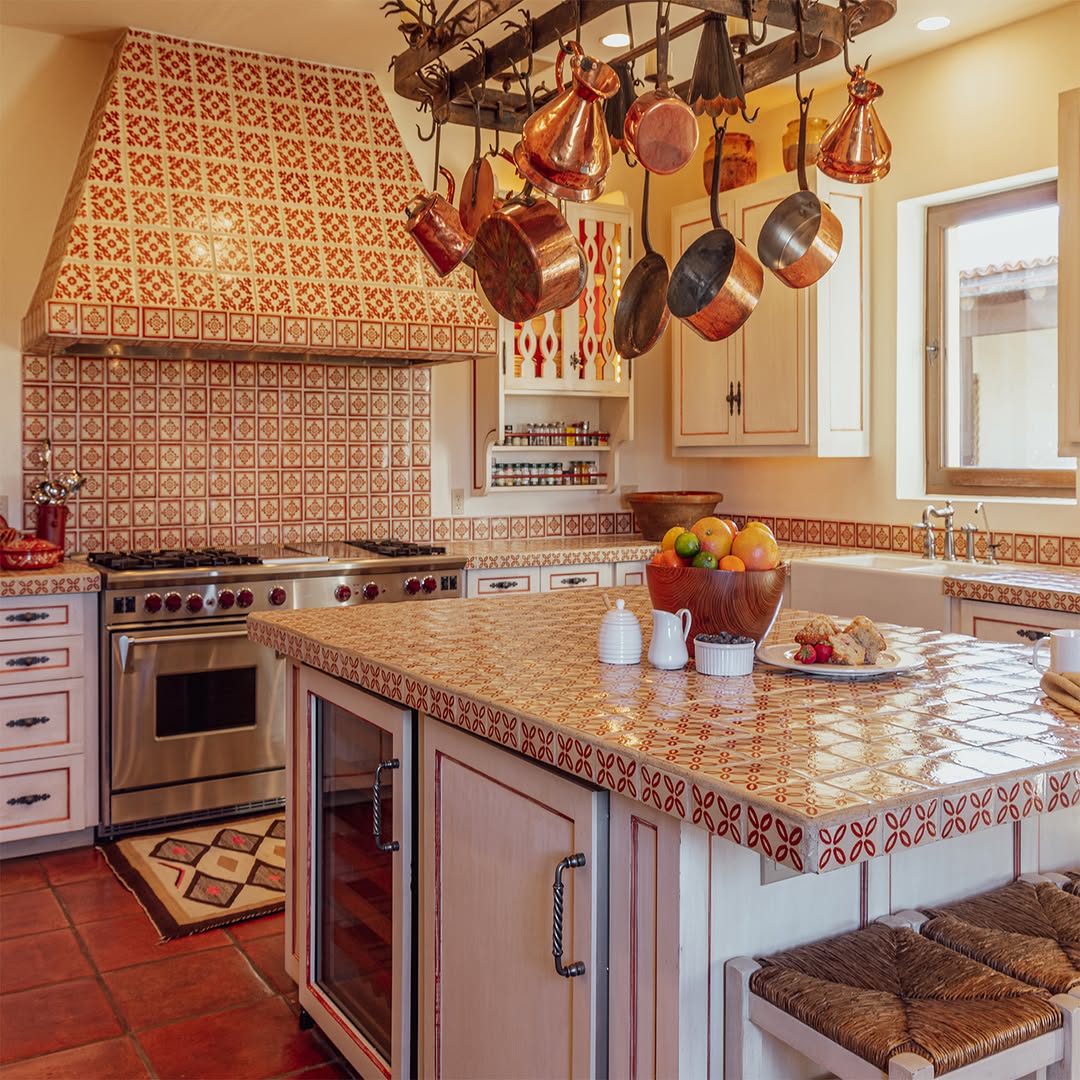 A mexican hacienda kitchen with patterned tile surfaces, a central island, stainless steel stove, hanging copper pots, and a bowl of fruit on the counter.