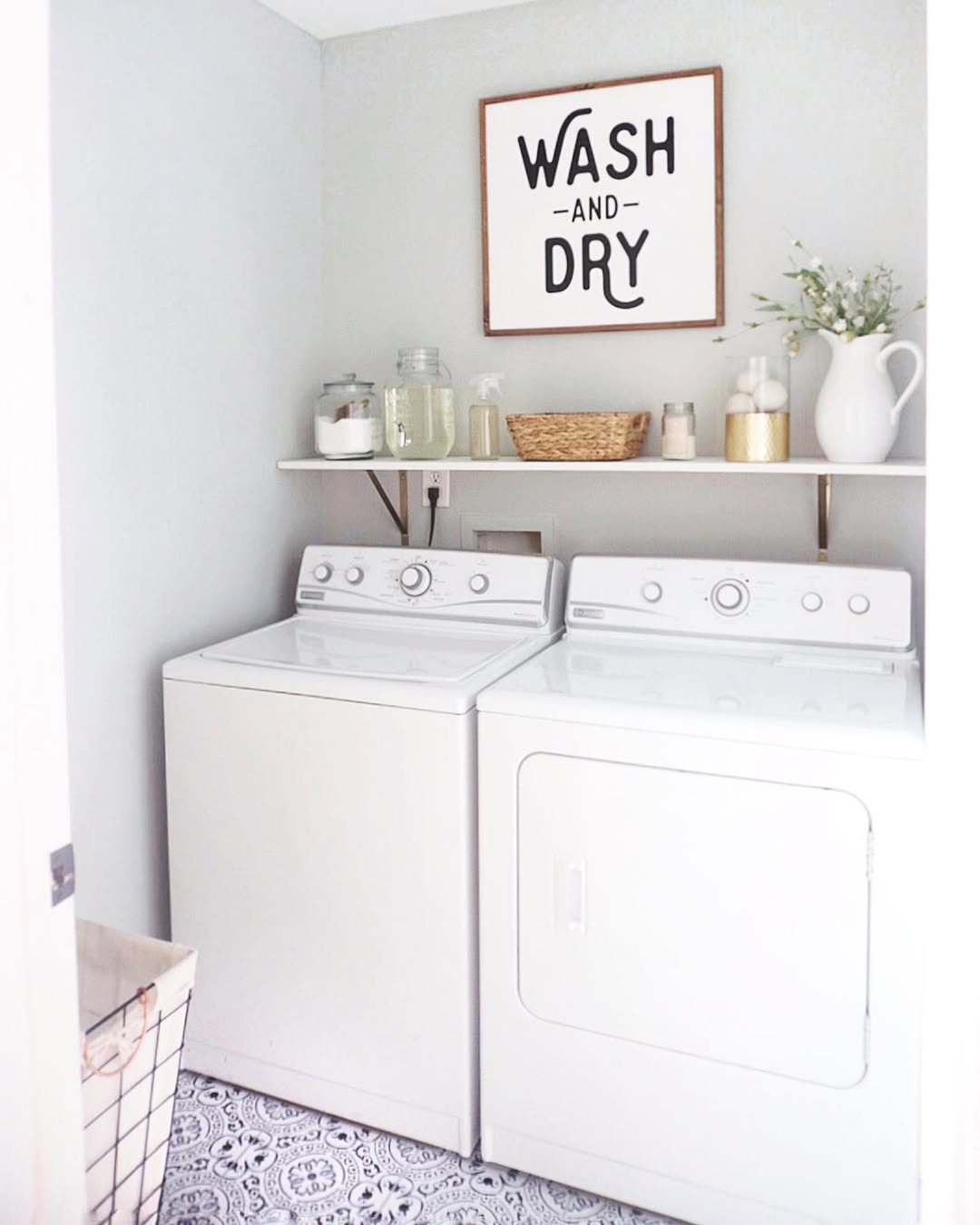 A small laundry room with a washing machine and dryer, a shelf holding jars and decor, a laundry basket, and a framed