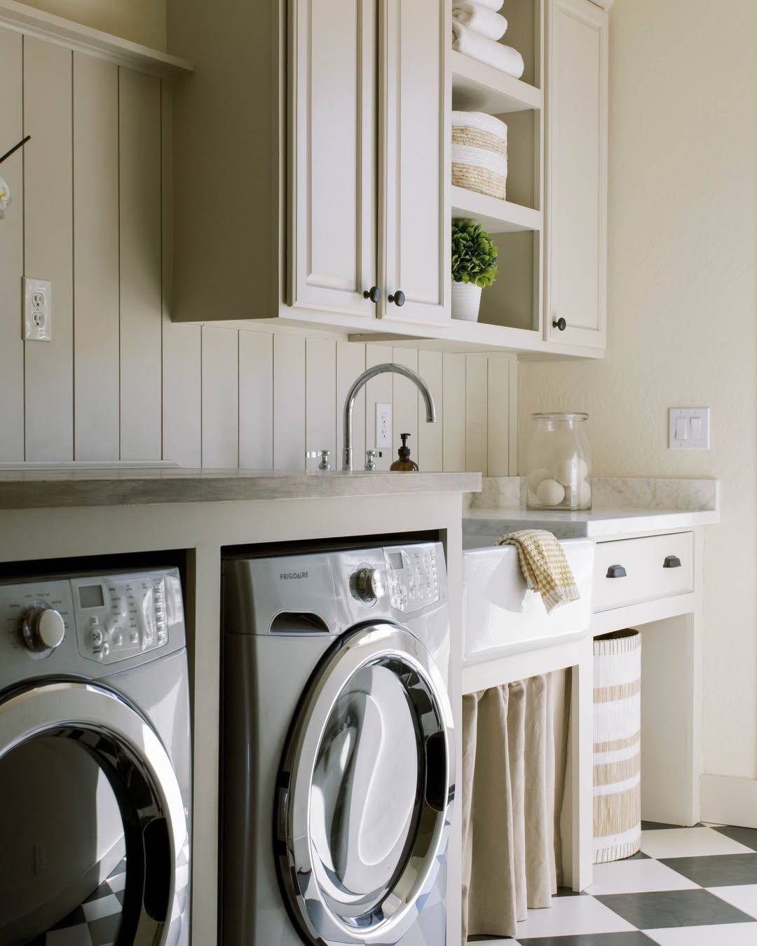 A Boho laundry room with a front-loading washer and dryer, white cabinetry, a sink, open shelves with boho decor, and a black-and-white checkered floor.