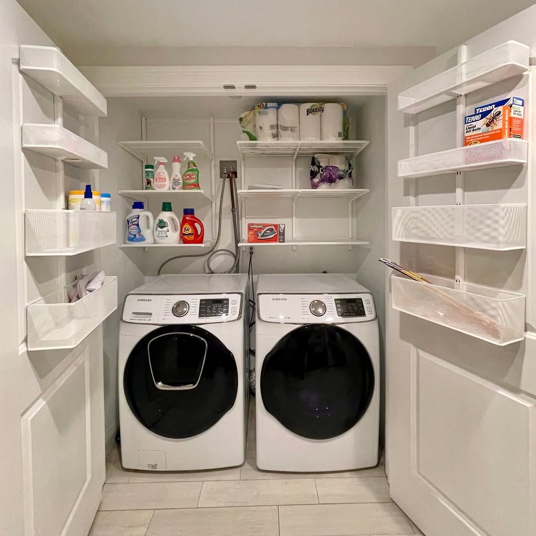 A small laundry room with a front-loading washer and dryer. Shelves on the sides neatly hold detergent, cleaning supplies, and paper towels, while upper shelves keep extra supplies and a basket organized.