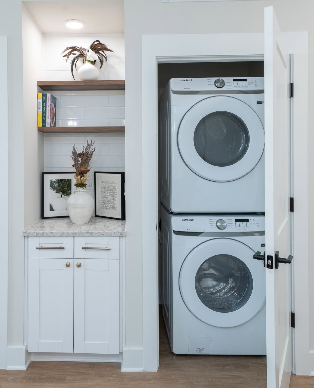 A stacked laundry room features a washer and dryer next to white cabinets, with open shelves displaying decor, books, and a vase with dried plants.