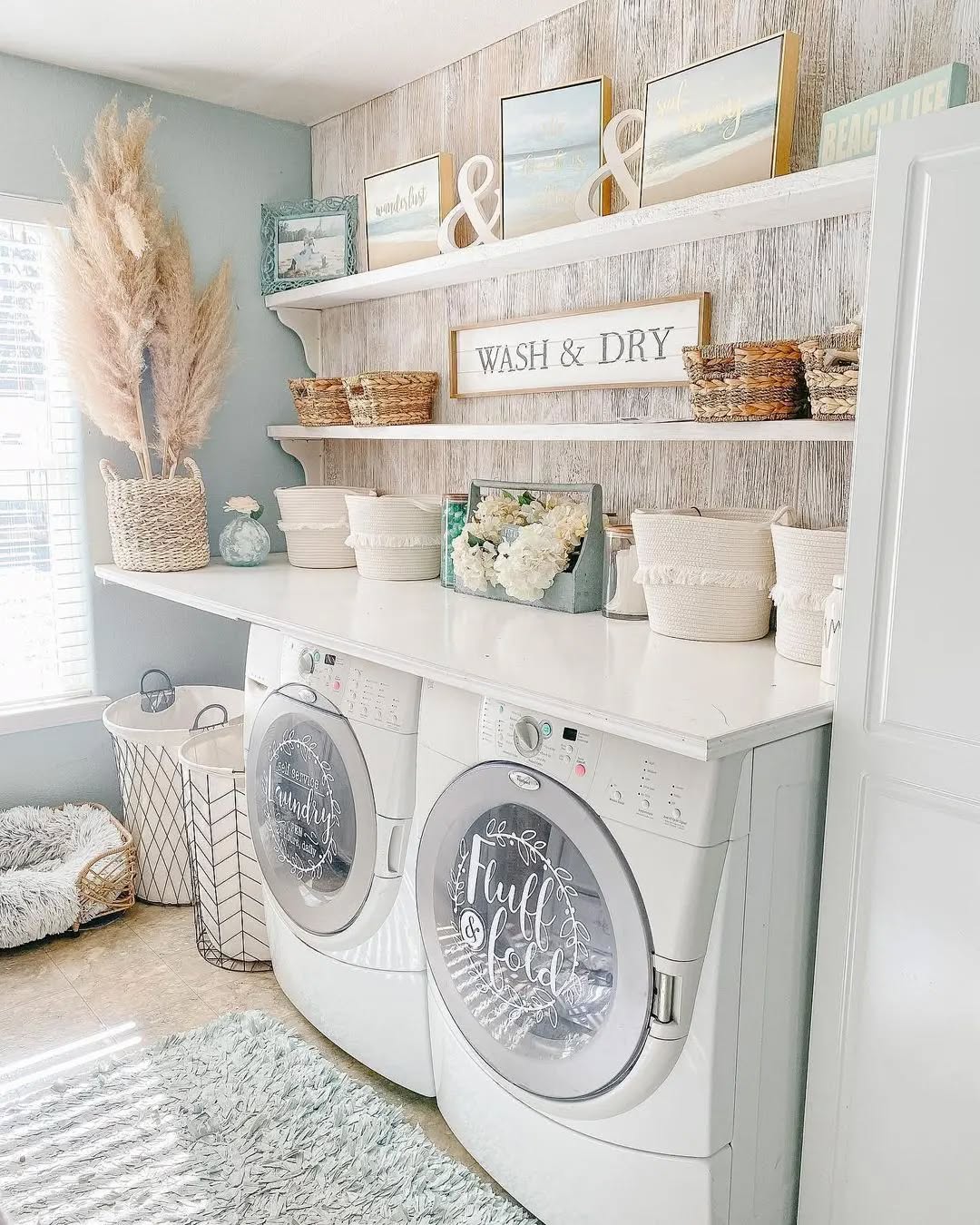 A bright, organized boho laundry room with a washer and dryer, baskets, decorative signs, shelving, and neutral-toned decor. Pampas grass and a rug add texture to this charming laundry room decor.