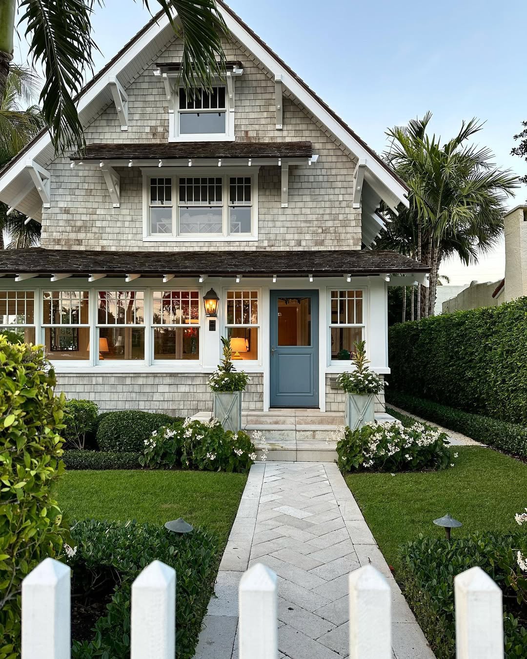 A two-story house with gray shingles, white trim, and a blue front door showcases a coastal cottage exterior, surrounded by manicured hedges and a stone path, viewed through a white picket fence—perfect inspiration for design ideas 2025.