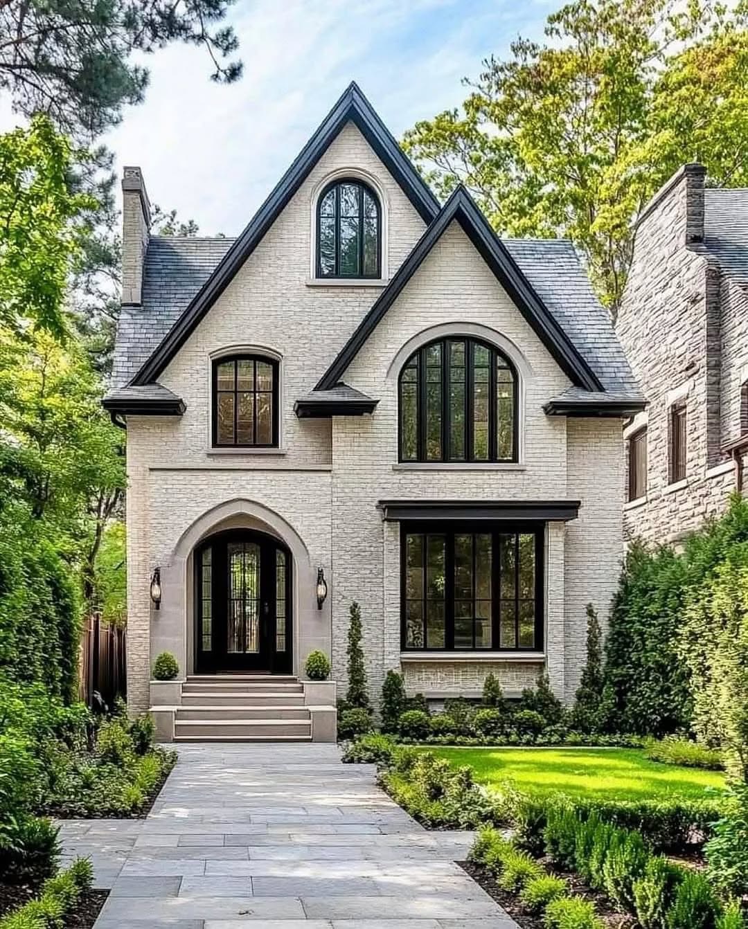 A two-story brick house with arched windows and a steep roof showcases charming exterior design ideas, surrounded by trimmed shrubs, a lawn, and a paved walkway leading to the front entrance.