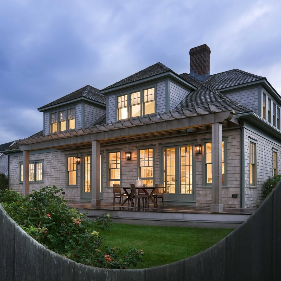 Two-story house with gray shingles, green trim, and a covered porch exudes coastal cottage exterior design. Outdoor dining table and chairs are on the porch; lights are on inside. Lush lawn and rose bush in front.