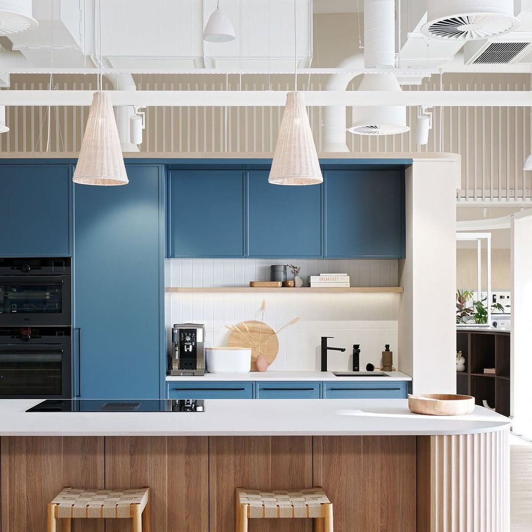 Modern kitchen with blue cabinets, wood accents, a white countertop island, pendant lights, and built-in appliances—showcasing one of the top kitchen trends 2025 for stunning kitchens and fresh kitchen color ideas.