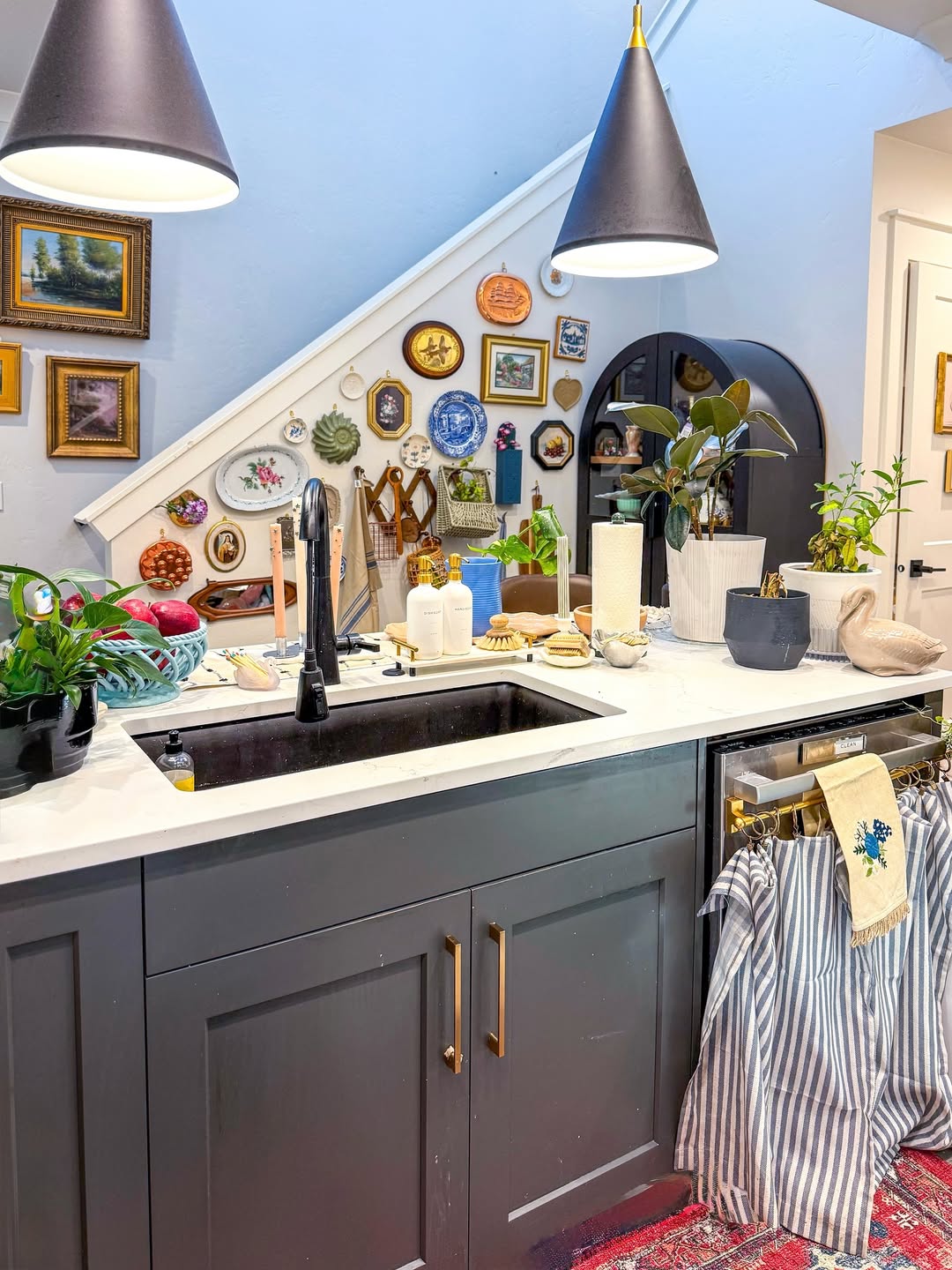 A kitchen with a black sink, modern pendant lights, and charming cottagecore kitchen decor. Plants, dishes, and utensils line the counter, while framed art pieces and plates decorate the wall under a staircase.
