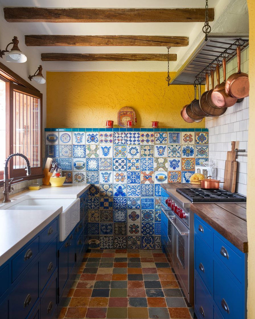 A small kitchen with bold color blue cabinets, a colorful patterned tile backsplash, wooden beams, hanging copper pots, and a multicolored tile floor—perfect inspiration for Mexican Hacienda kitchen design ideas.