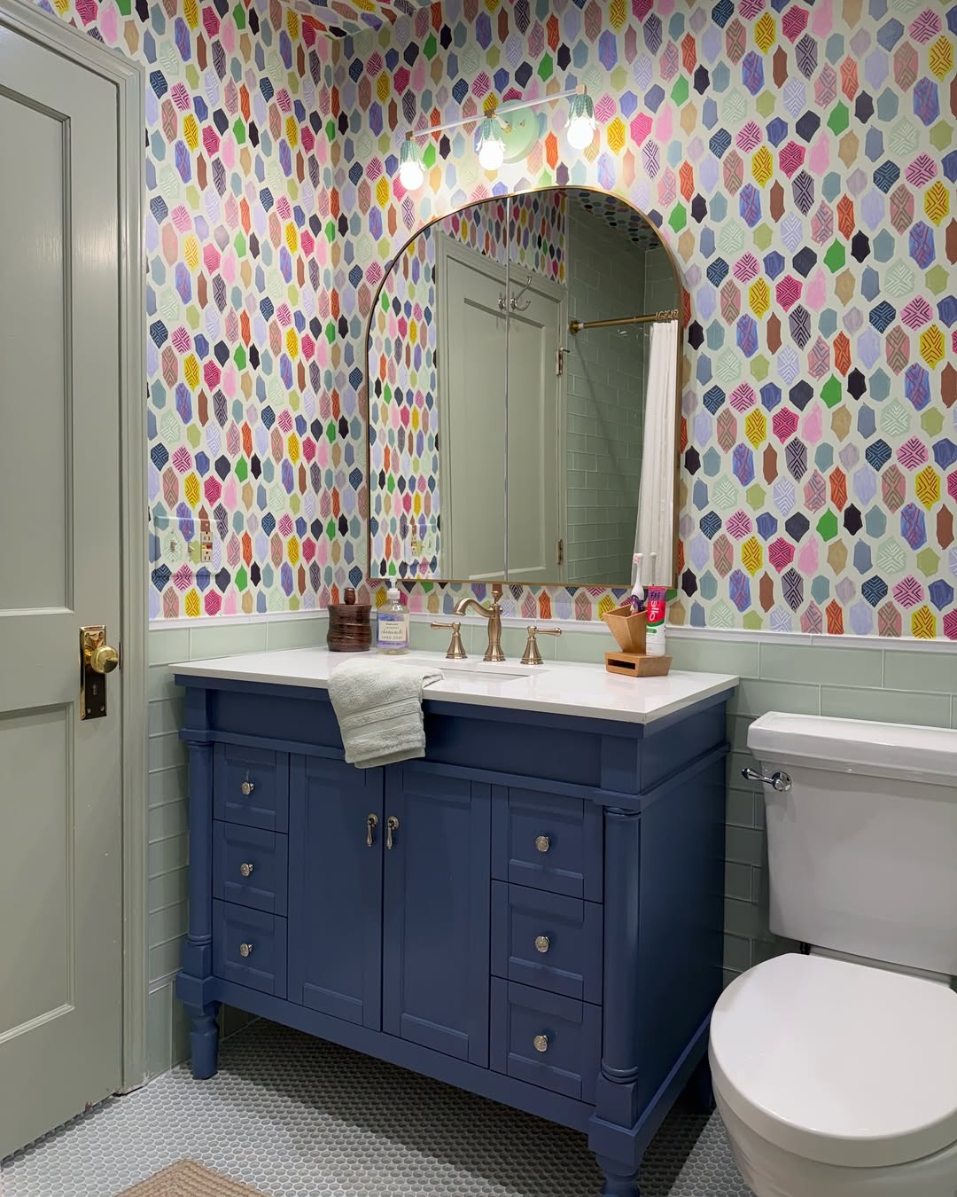 A playful kids bathroom featuring colorful geometric wallpaper, a blue vanity with a sink, a mirror with two lights, and a white toilet.