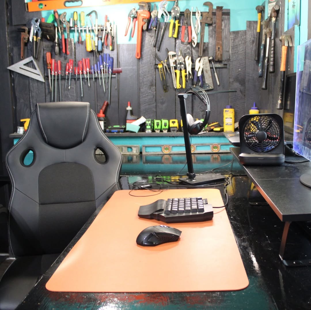 A black desk chair sits at a desk with a mouse, split keyboard, and fan—perfect for a home office workshop; behind the desk, a wall displays various hand tools hanging on hooks.