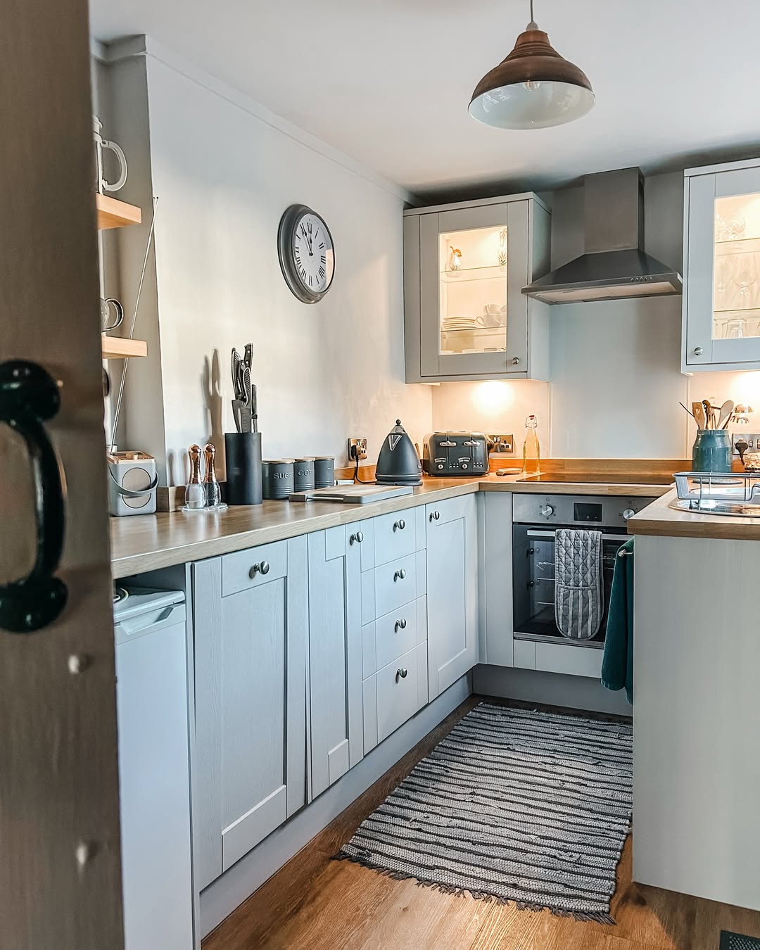 Modern kitchen with light gray cabinets, wooden countertops, shelves with utensils, a stove, oven, and a striped rug on a wooden floor. A wall clock above the countertop adds charm—perfect inspiration for farmhouse kitchen ideas.