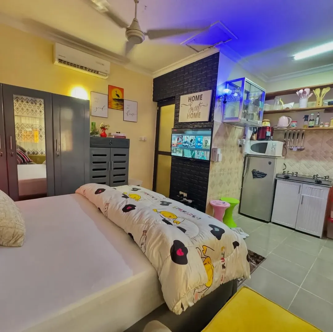 A compact studio apartment featuring a bed, wardrobe, TV, small kitchen area, wall shelves, and bright lighting—decorated with wall art and patterned bedding for smart Studio Apartment Ideas.