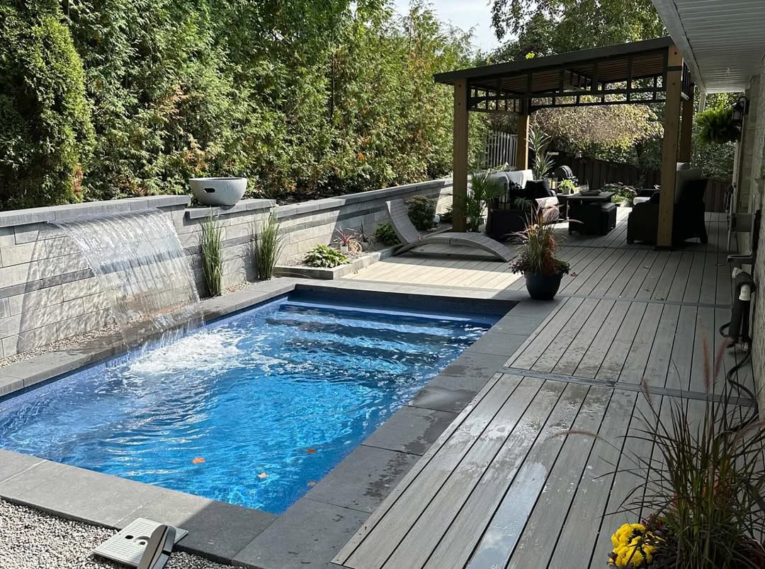 A small backyard pool with a waterfall feature is surrounded by gray decking, lush plants, and a covered outdoor seating area—perfect inspiration for cool pool ideas.