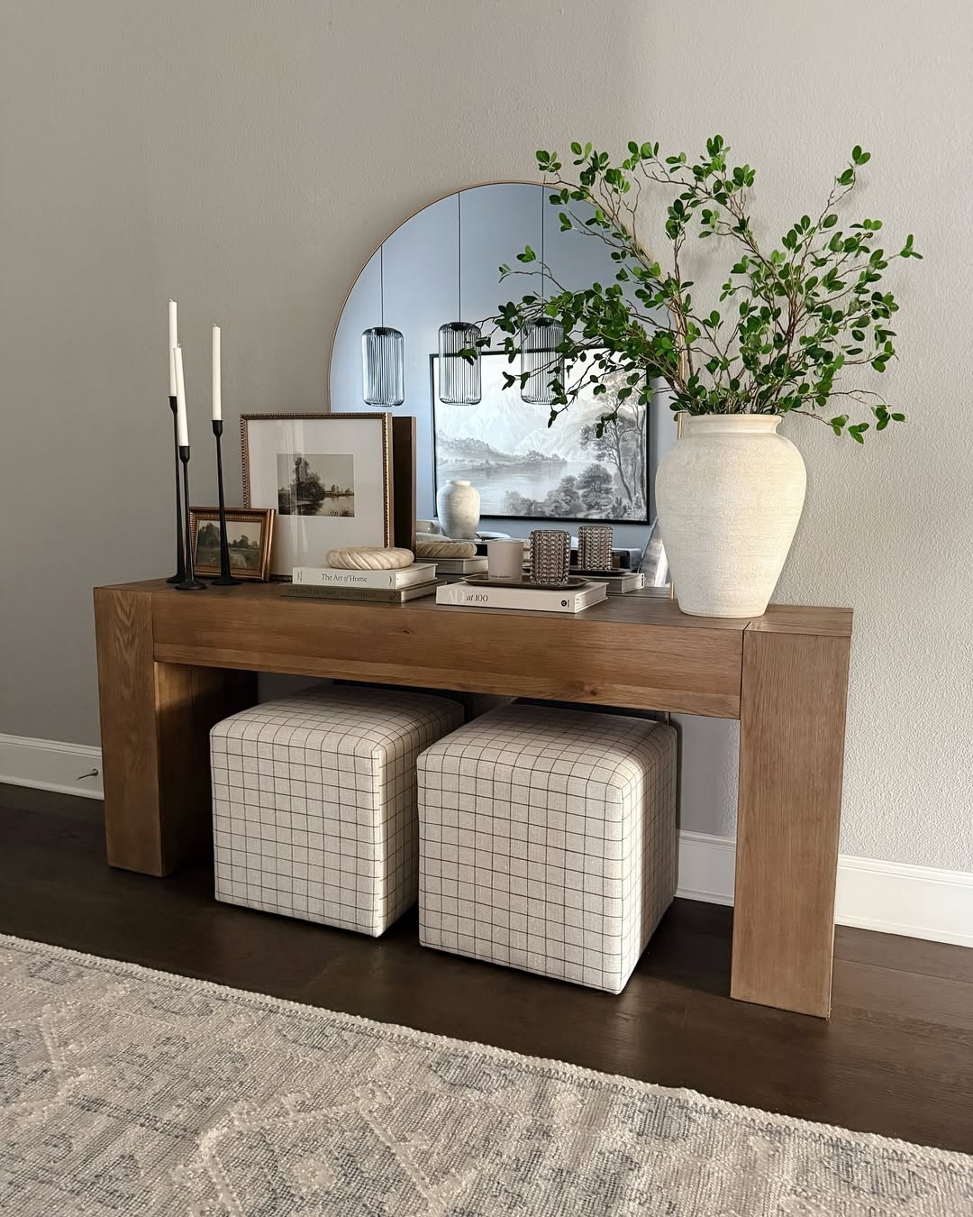 A wooden console table features stylish console table decor: a large round mirror, white vase with green branches, books, framed art, candles, and two plaid ottomans underneath. Perfect for those seeking fresh console table ideas.