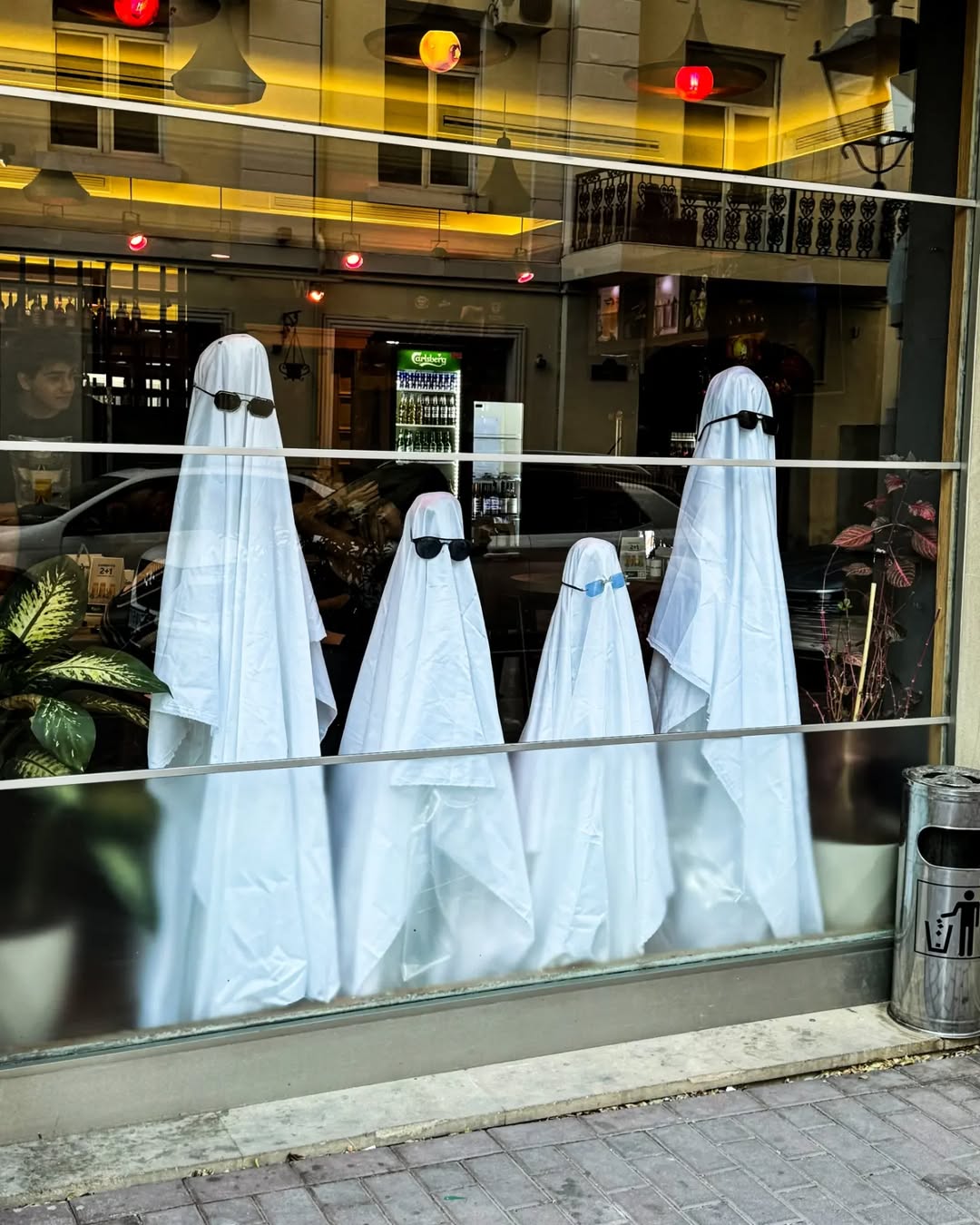 Four ghost figures made from white sheets and sunglasses stand behind a glass window in a restaurant or cafe, serving as creative Halloween window decorations with plants and indoor lighting in the background.