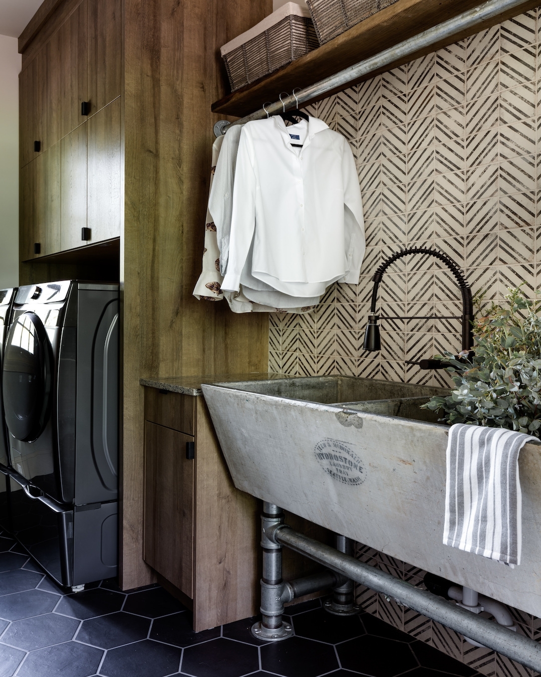 A laundry room with a washer and dryer, a large stone utility sink, wood cabinets, a shirt hanging above the sink, and patterned tile backsplash offers practical utility room style and inspiration for laundry room decor.