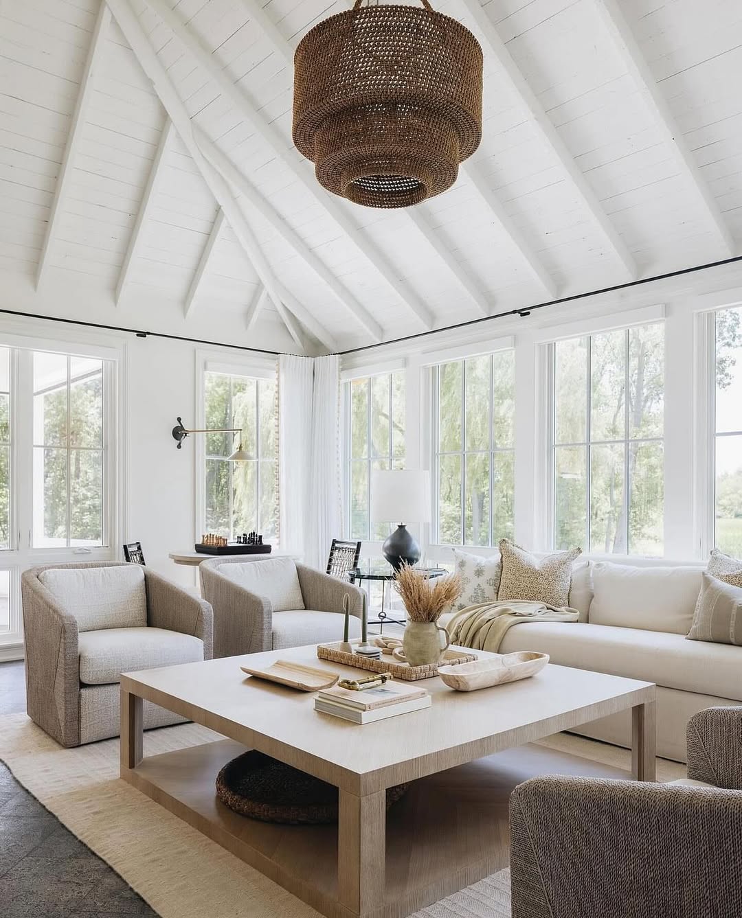 Bright organic modern living room with a vaulted white ceiling, large windows, neutral-toned furniture, a central wooden coffee table, and a woven hanging light fixture—perfect inspiration for modern living room ideas.