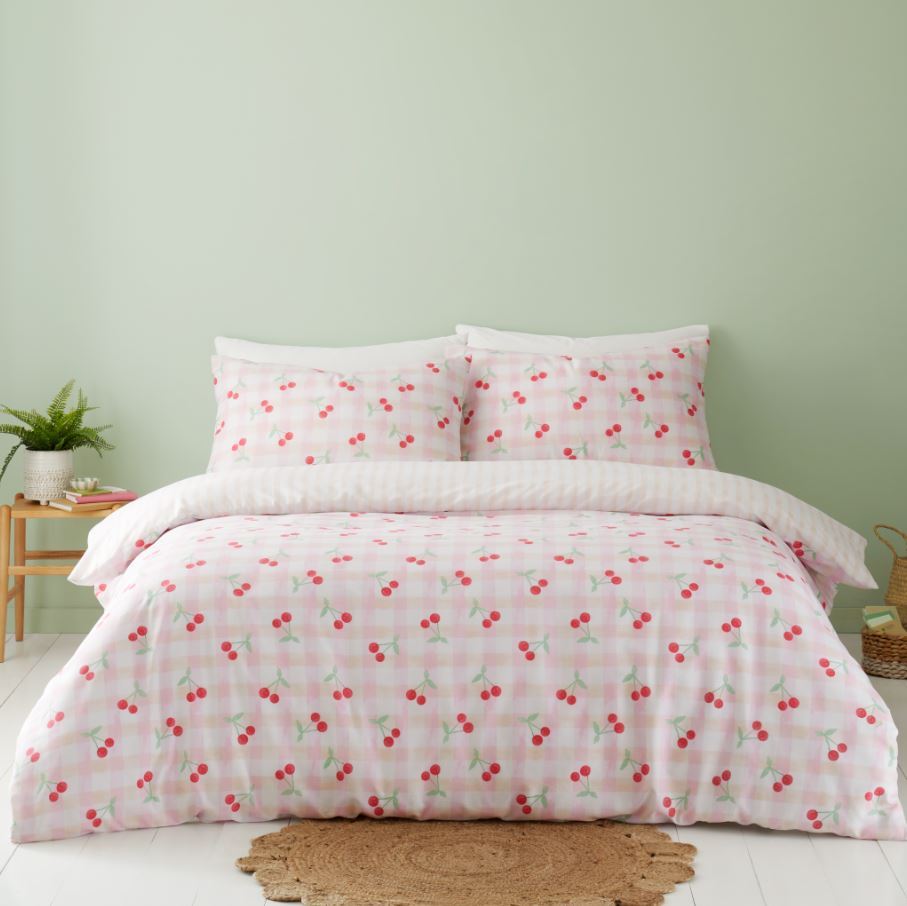 A neatly made bed with cherry-patterned pink and white bedding, two matching pillows, a round woven rug, and a small wooden side table with a potted plant—perfect inspiration for charming cherry bedroom ideas and cozy bedroom decor.