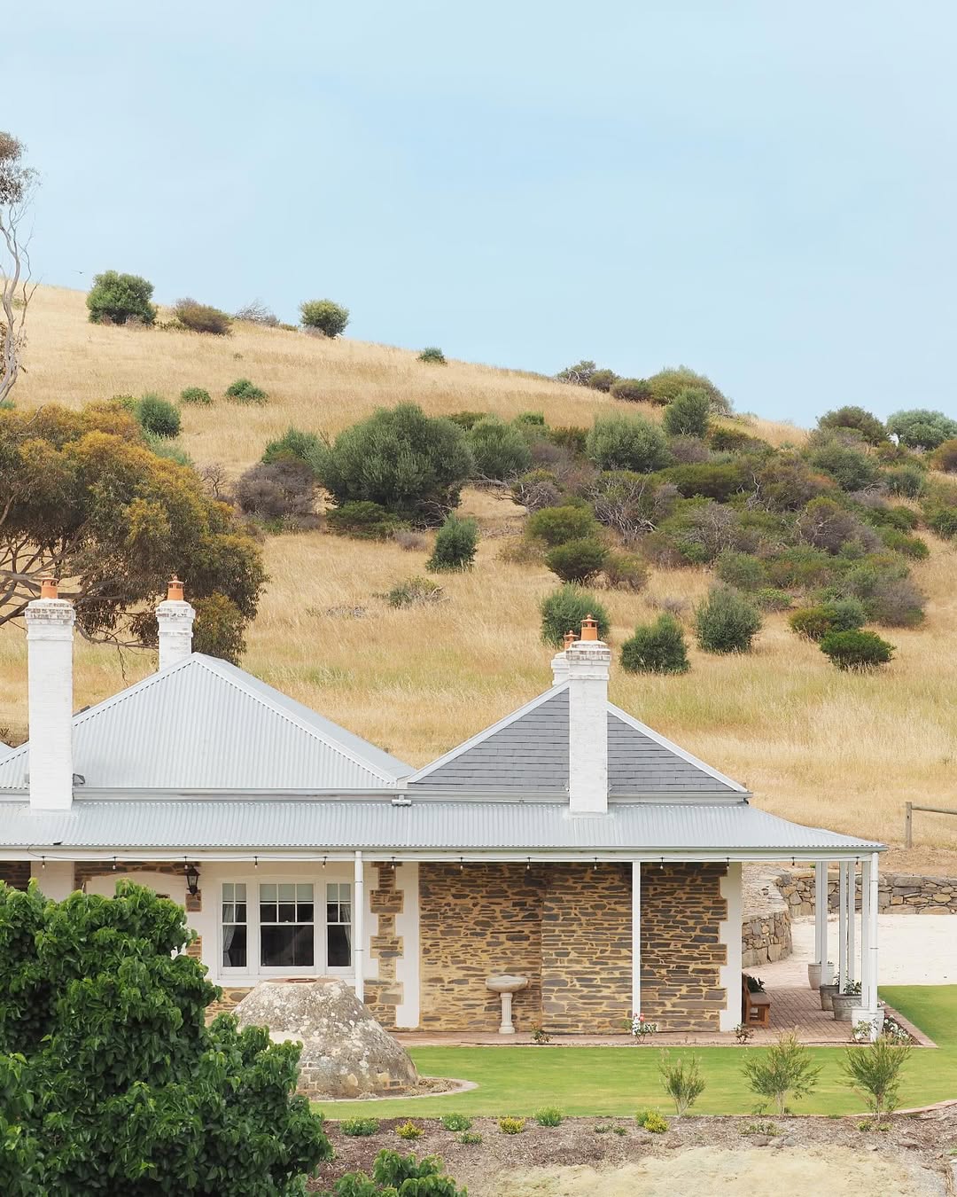 A single-story stone house with a gray metal roof and white chimneys sits in front of a grassy hillside, offering ultra-stylish coastal cottage exterior vibes perfect for those seeking design ideas for 2025.