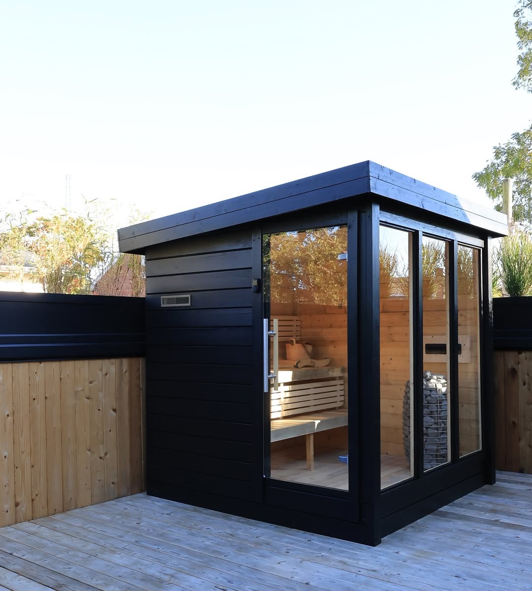 A modern black and wood-paneled outdoor sauna with large glass doors and windows, situated on a wooden deck in a fenced yard—perfect inspiration for stylish outdoor sauna designs.