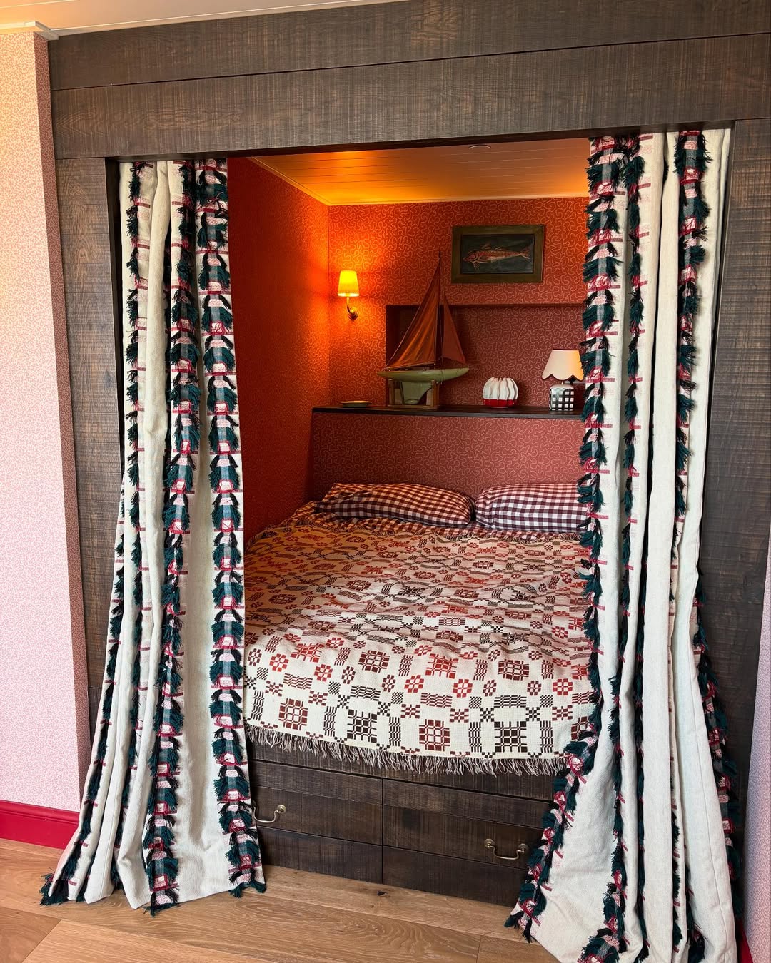 A cozy bed with checkered pillows and a patterned quilt is set in a wooden alcove, flanked by patterned curtains—perfect inspiration for small bedroom layout ideas. Nautical decor and a lamp rest on the shelf above the bed.