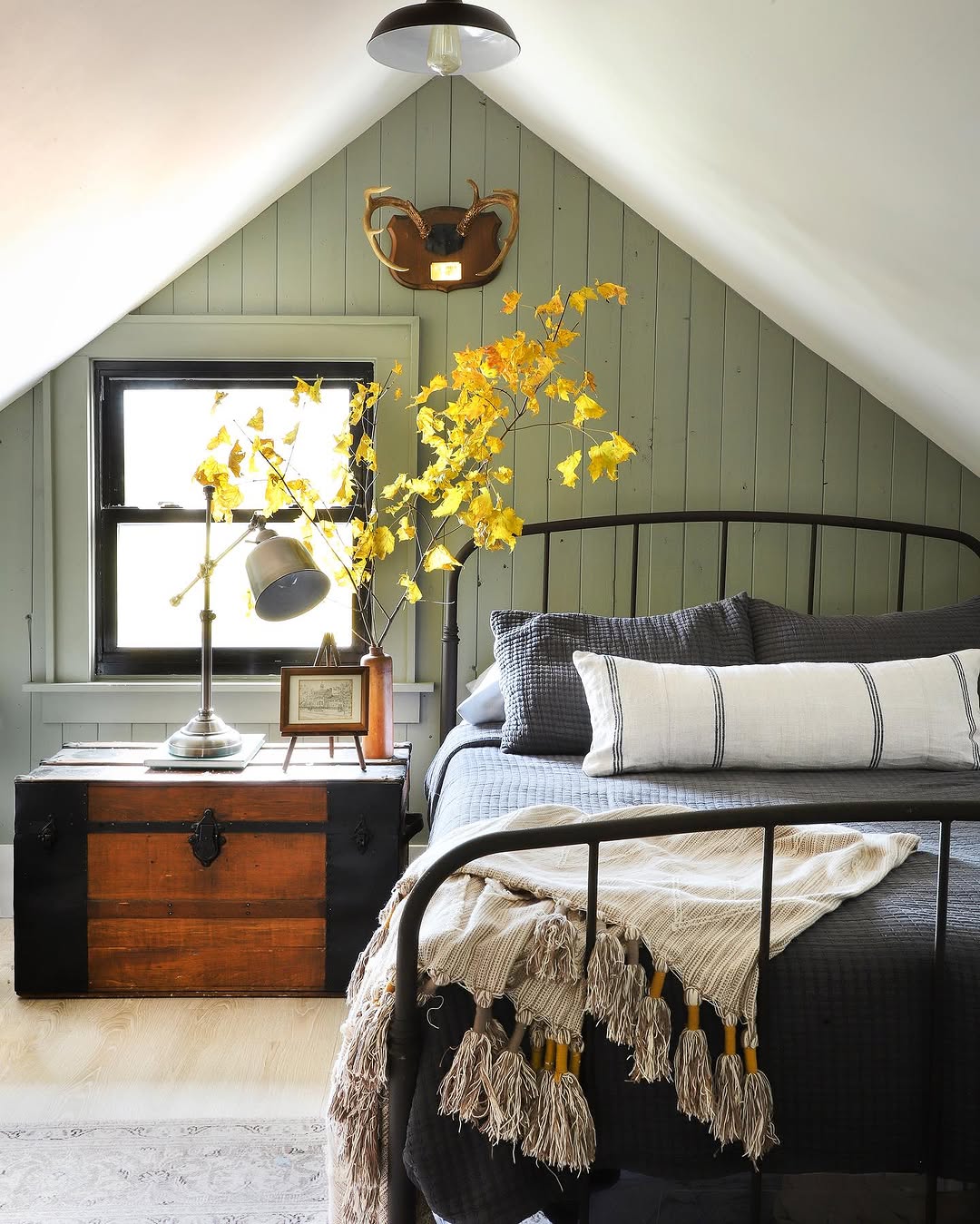 A cozy sage green farmhouse bedroom attic with a black metal bed, striped pillows, a gray tasseled blanket, wooden trunk, table lamps, and yellow flowers in a vase beneath the sloped ceiling.
