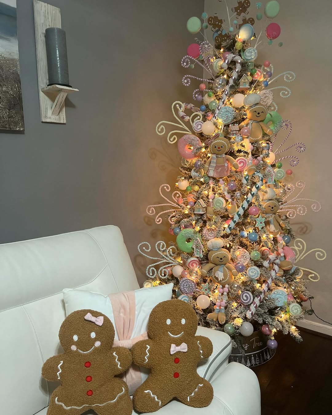 A Christmas tree adorned with candy and gingerbread ornaments stands beside a white couch with two gingerbread-shaped pillows, creating charming Gingerbread Decor.